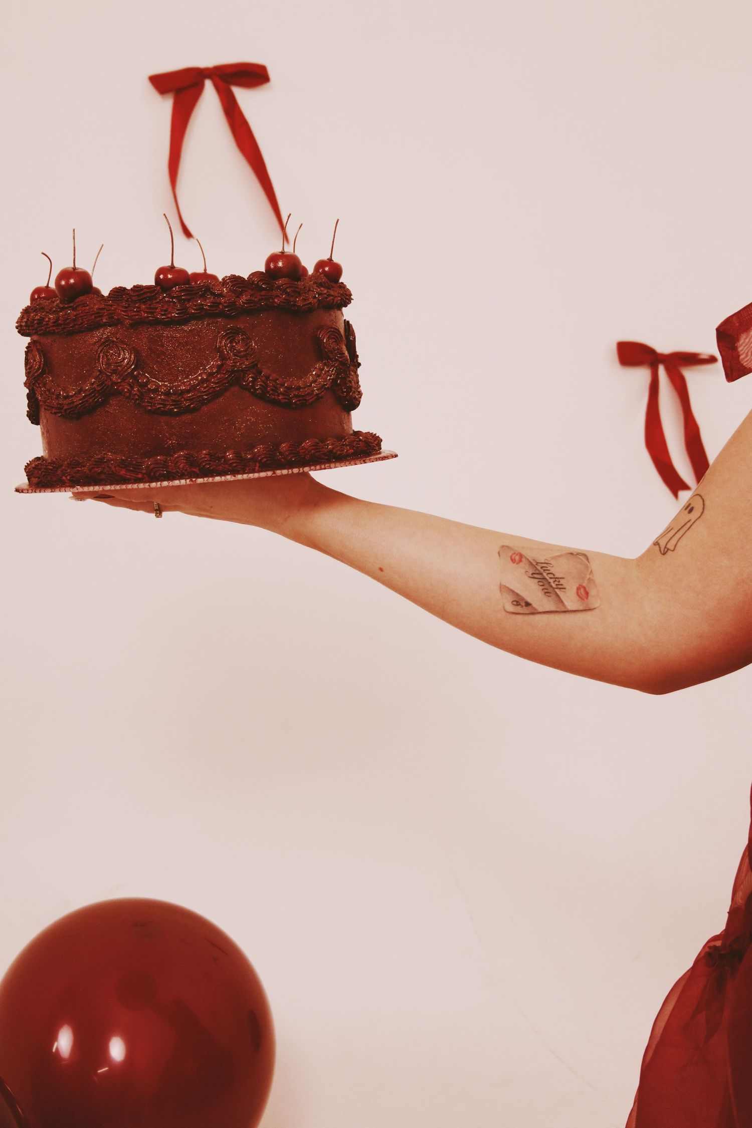 Person holding a decorated chocolate cake with cherries and red ribbon bows on a tray, with a red balloon below.
