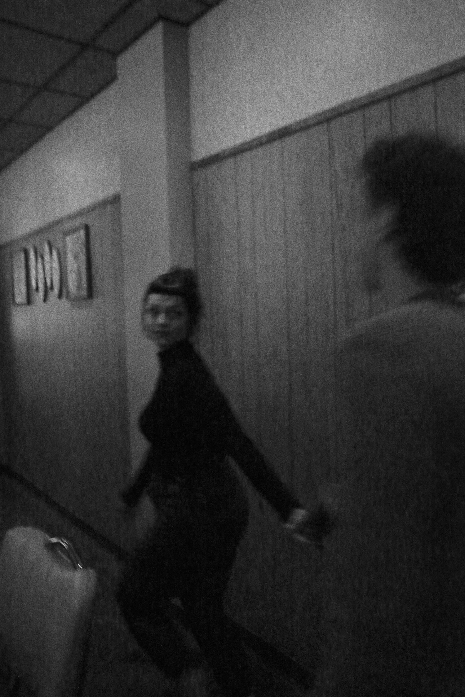 Black and white photo of a woman in a dark outfit walking down a hallway with wood-paneled walls.