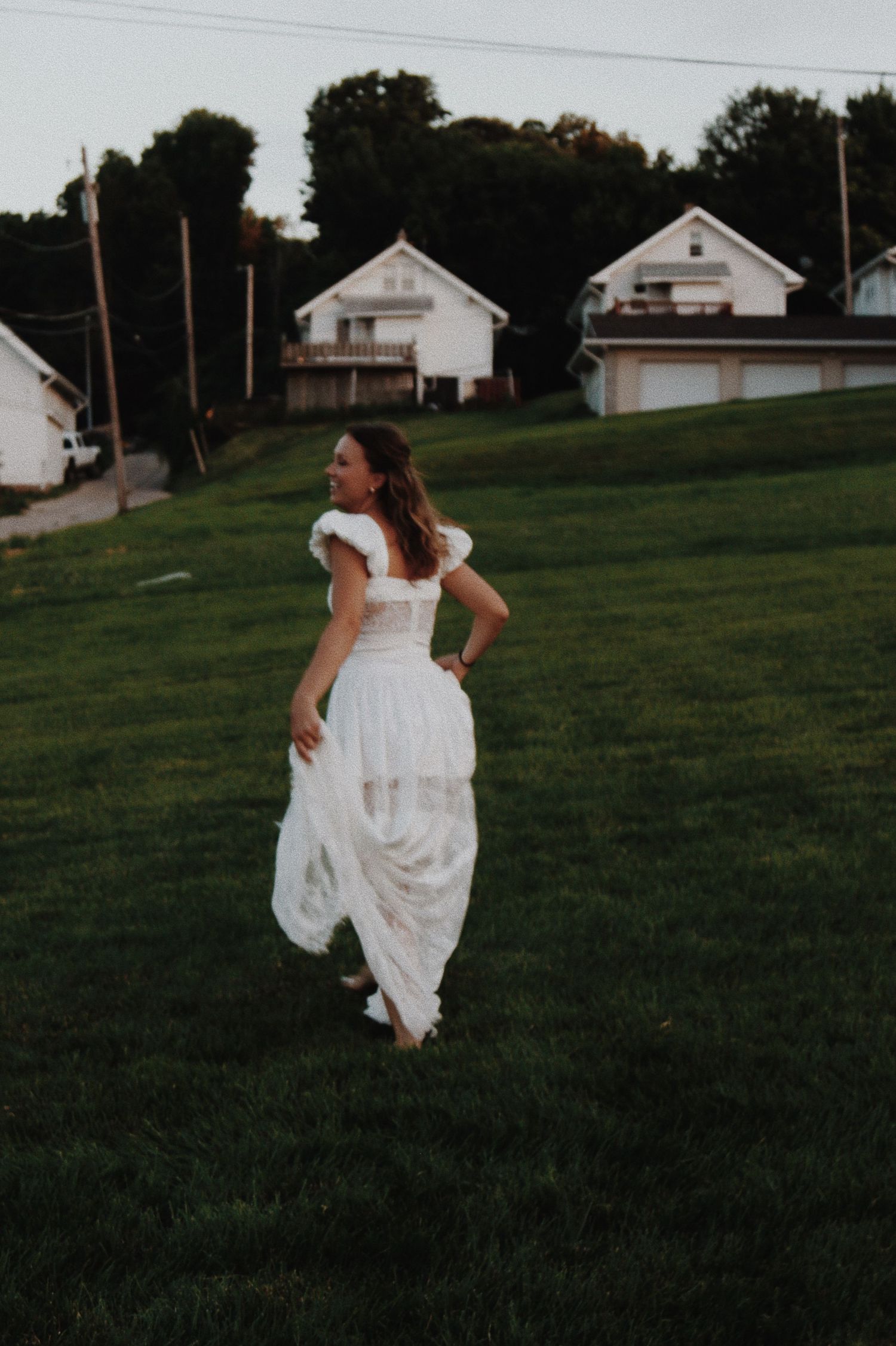 Woman in white lace wedding dress walking barefoot on green grass at dusk.