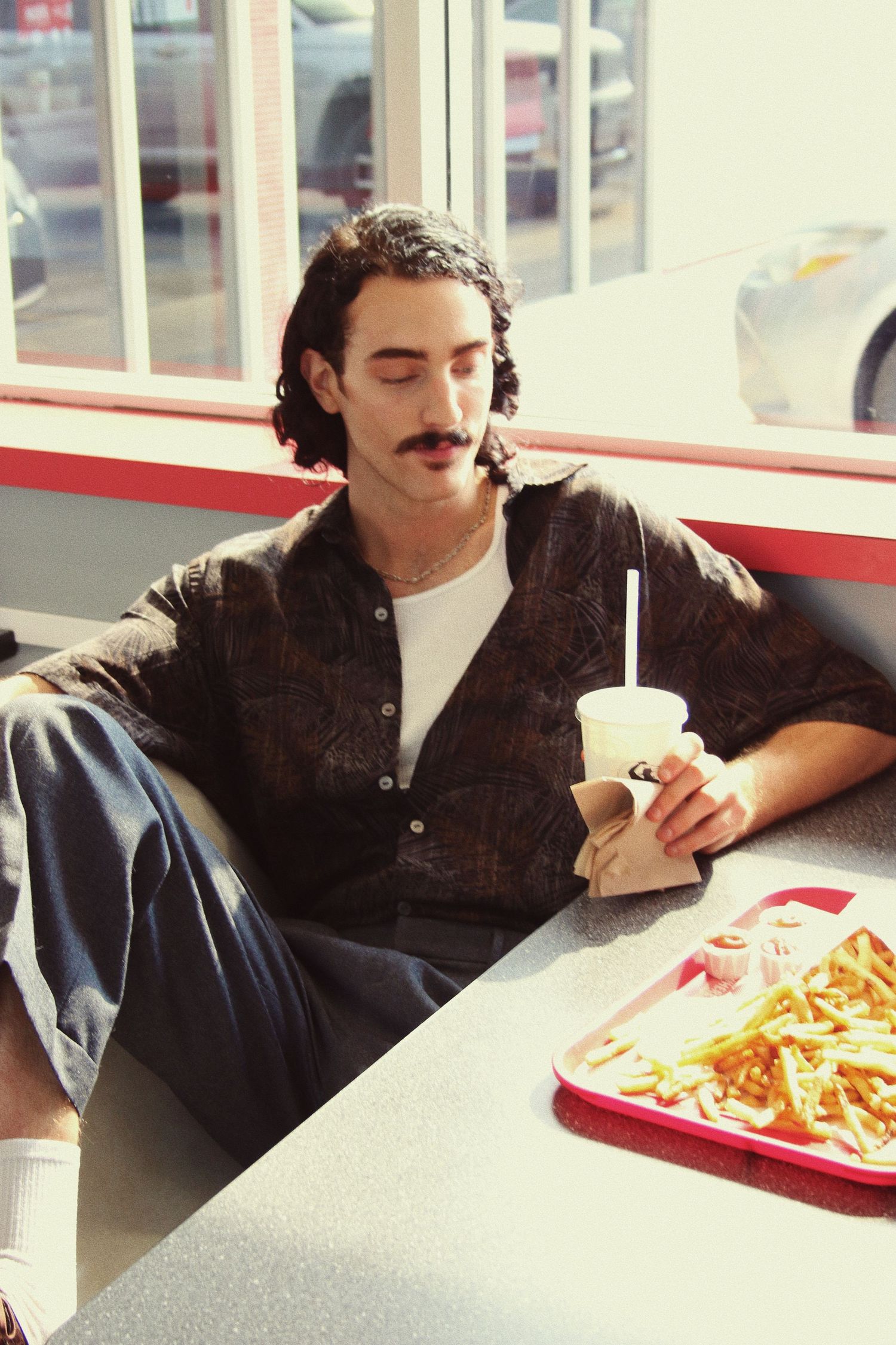 Young man with mustache sitting in fast food restaurant, holding a drink with fries on tray nearby.