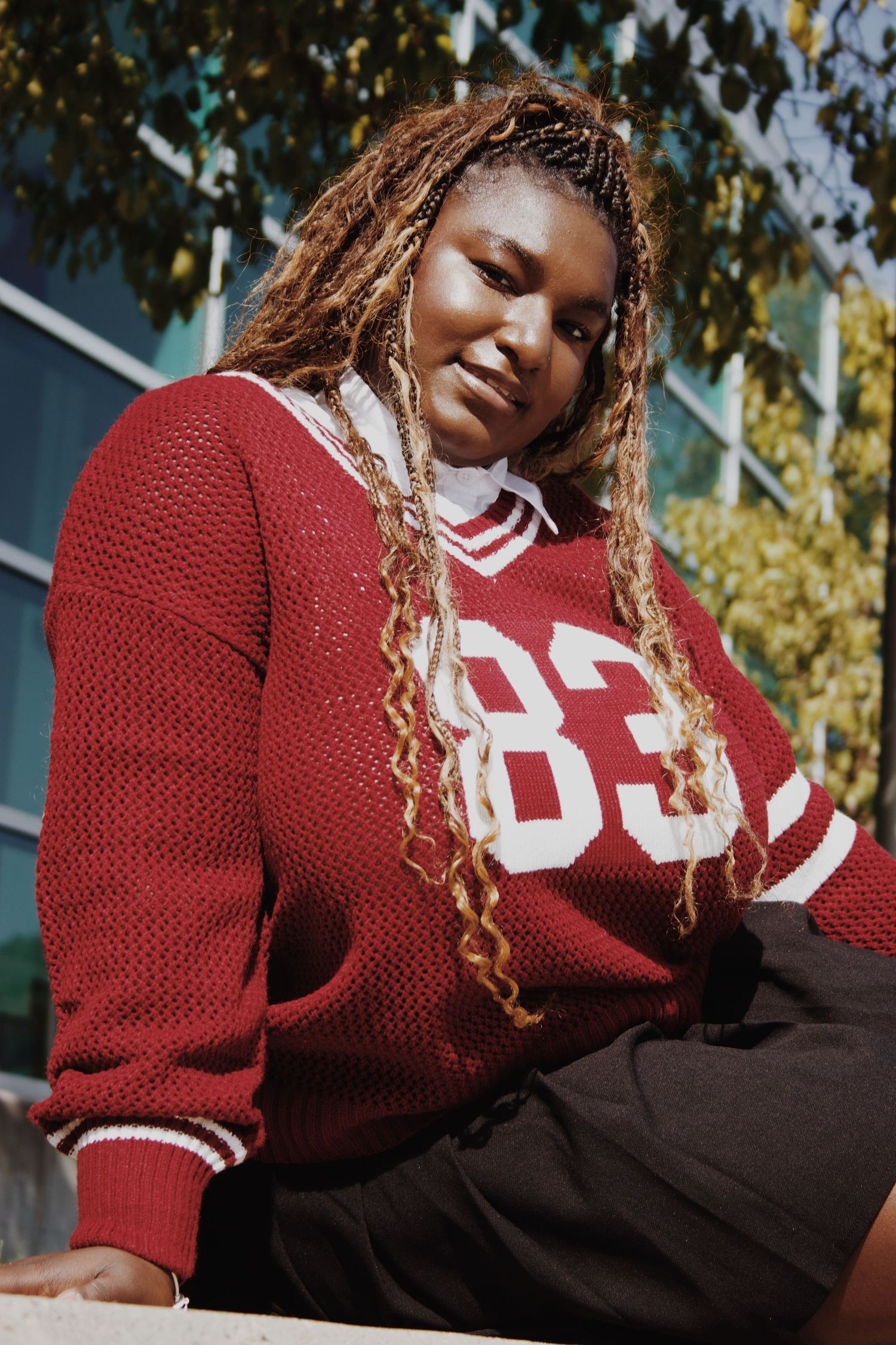 Young woman in red number 83 varsity sweater with braided hair poses outdoors in sunlight.