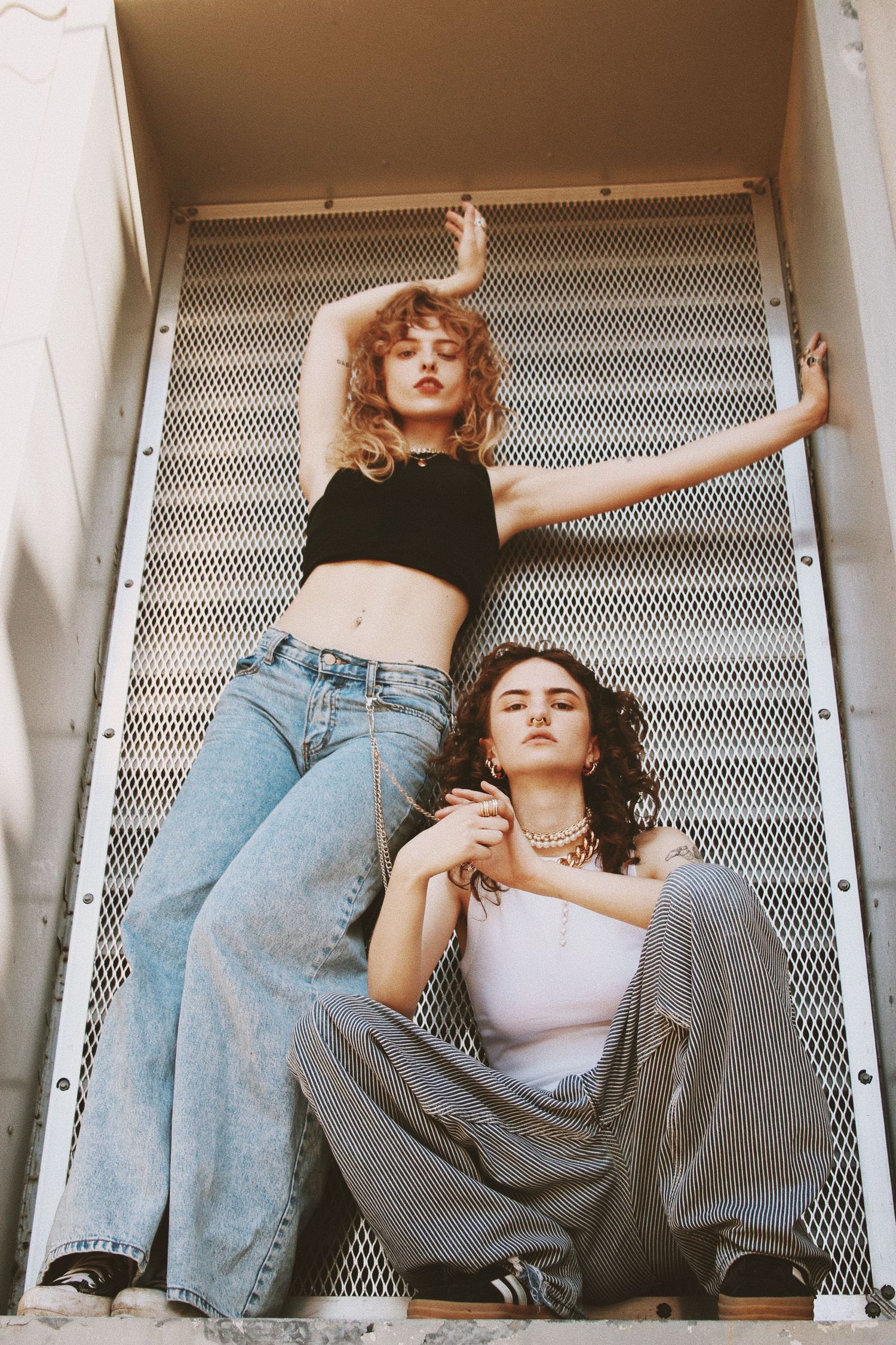 Two young women in wide-leg jeans pose against a metal grate backdrop in a trendy urban fashion shoot.