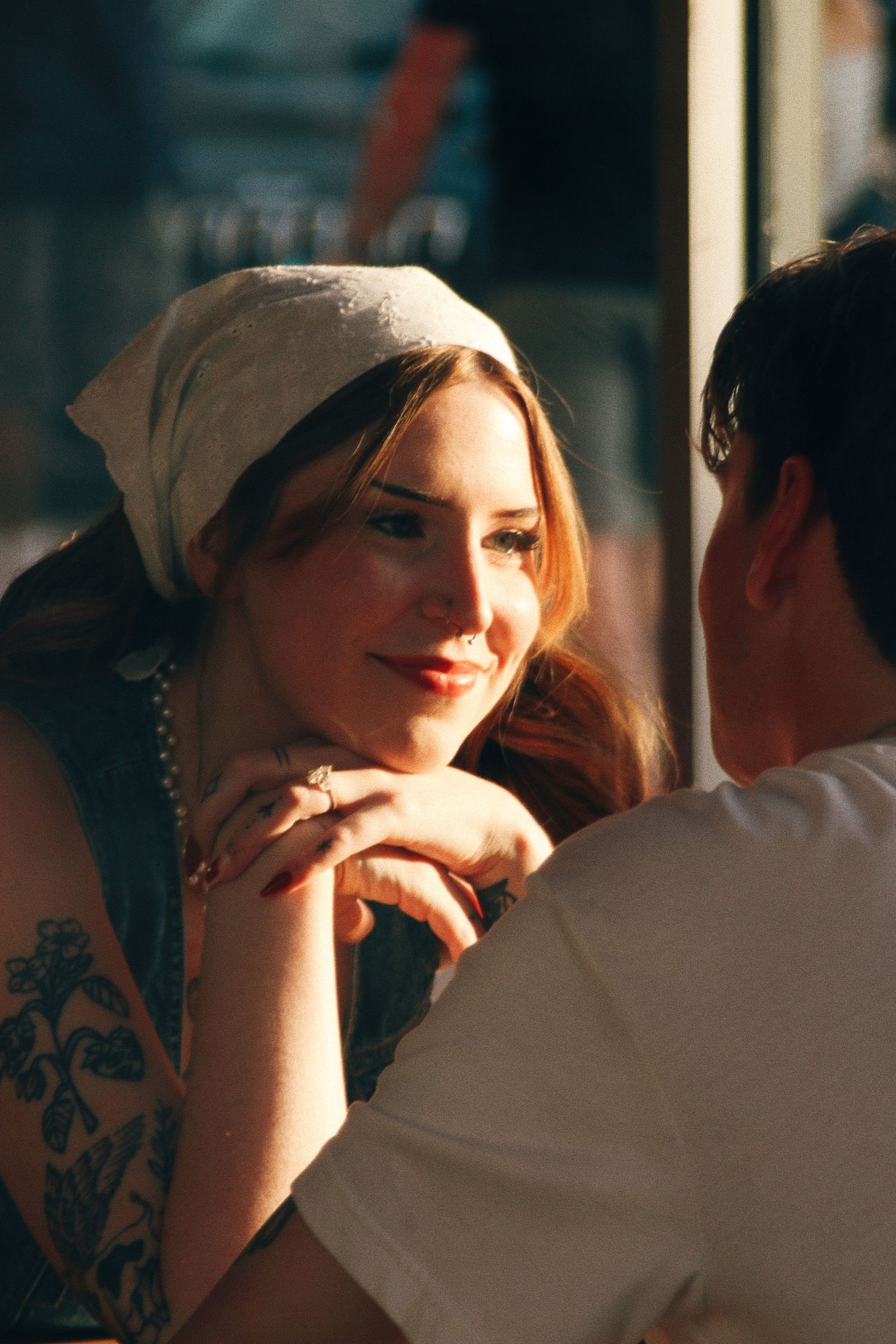 A smiling woman wearing a headscarf and floral tattoo chats with a man in warm golden sunlight.