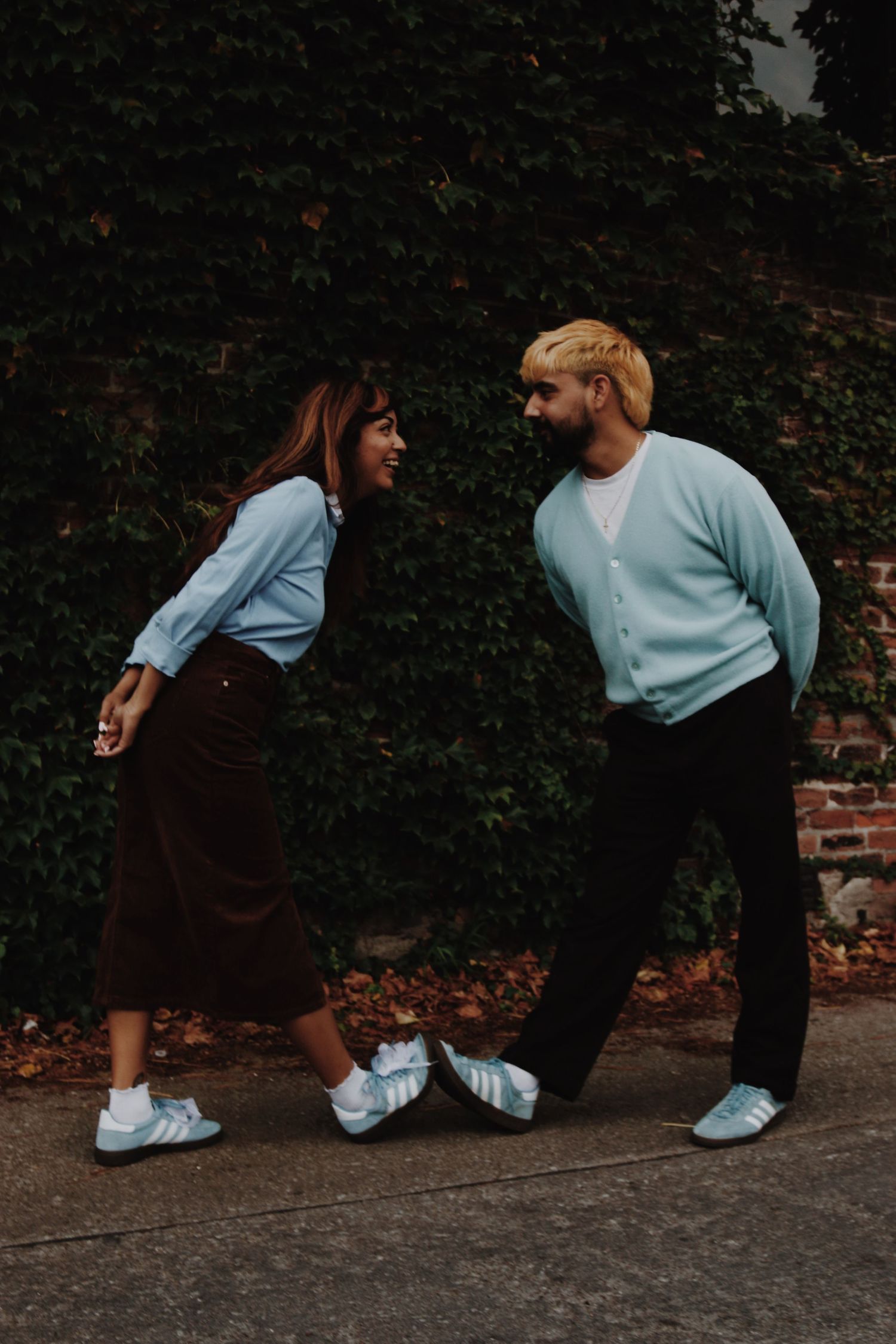 A smiling couple in matching light blue cardigans and sneakers playfully face each other on a sidewalk near an ivy-covered wall.