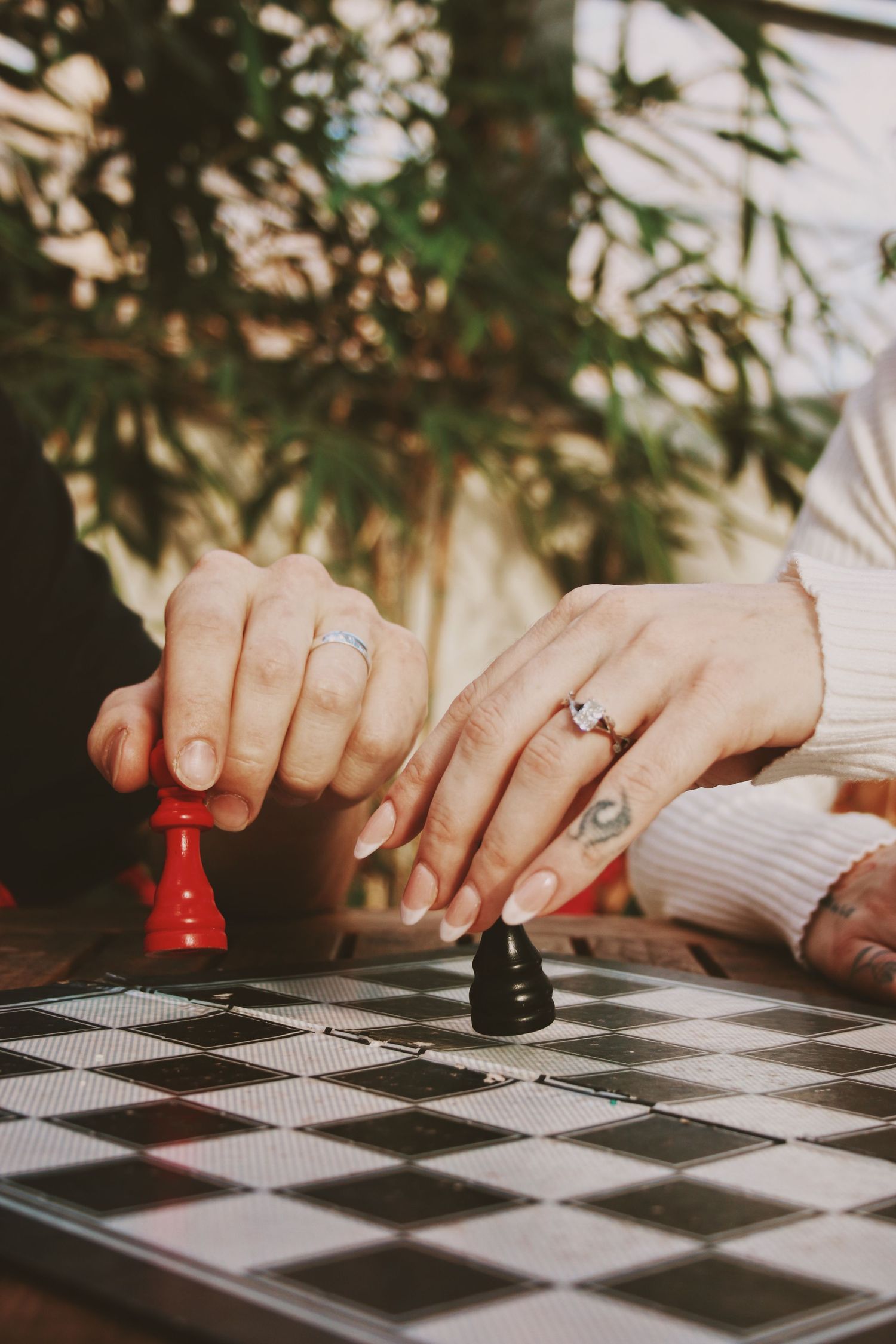 Two hands with rings move red and black chess pawns on a checkered board, with green plants in the background.