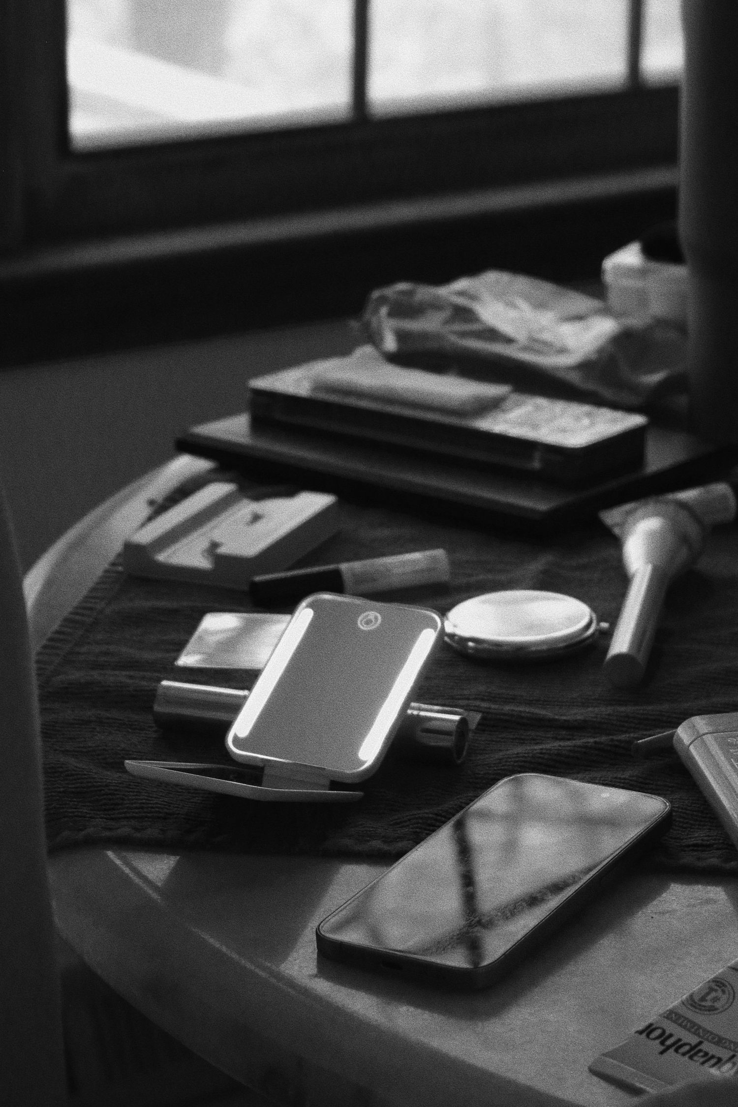 Black and white photo of makeup items and a smartphone scattered on a vanity table near a window.