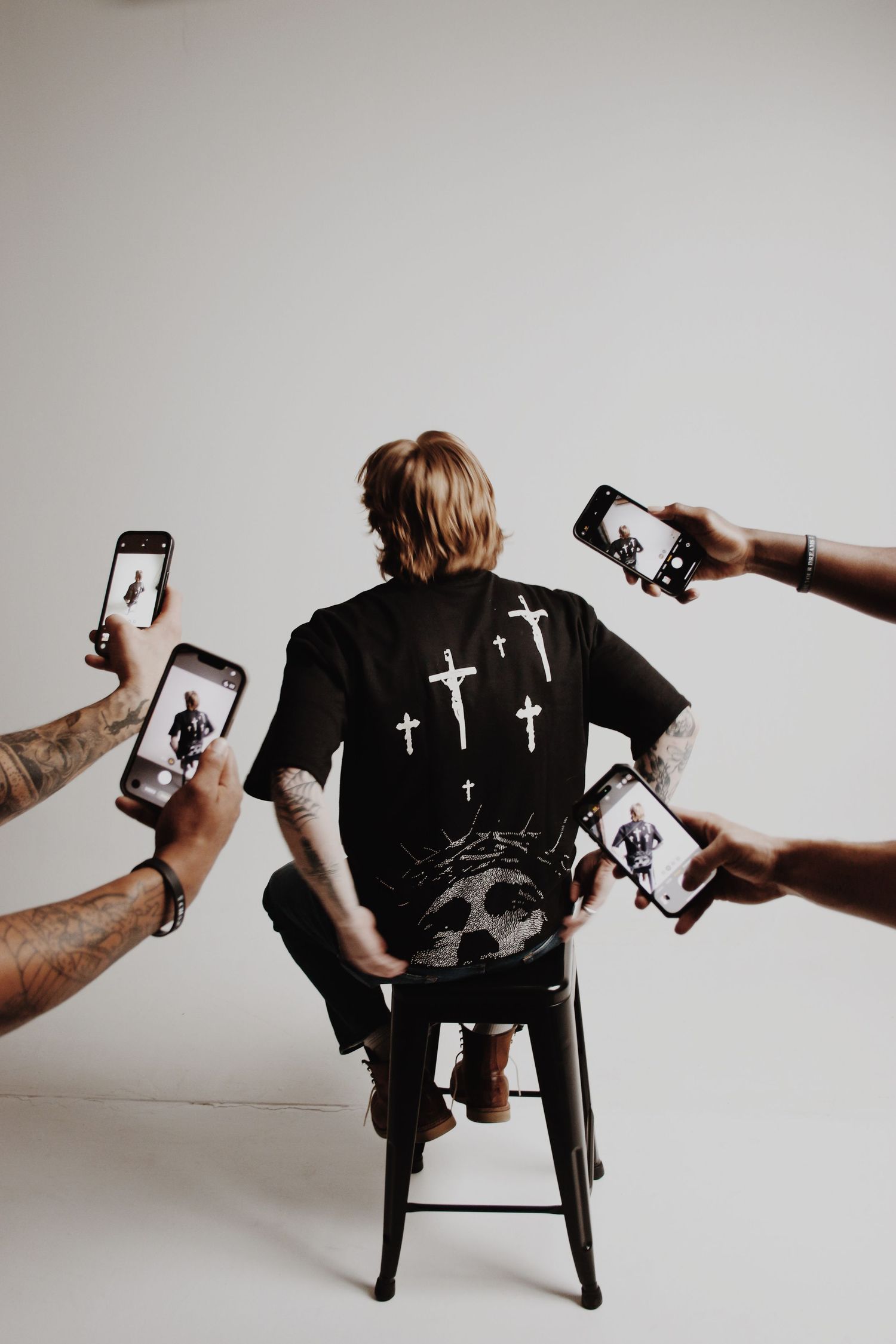 Person sitting on stool with back turned, wearing black cross graphic tee, surrounded by hands holding smartphones photographing them.