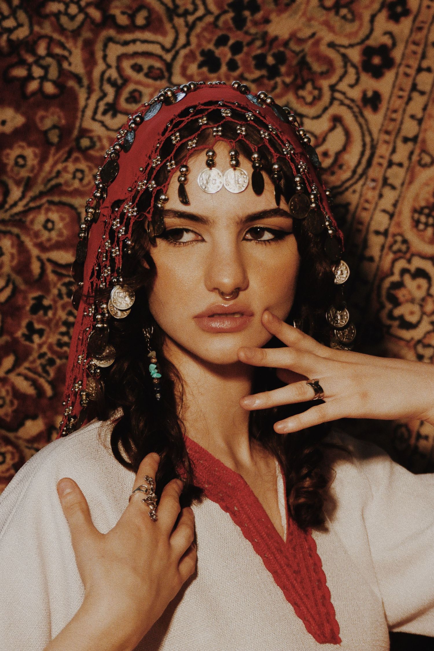 Woman wearing ornate beaded headpiece with coins and red scarf, posing against decorative tapestry background.