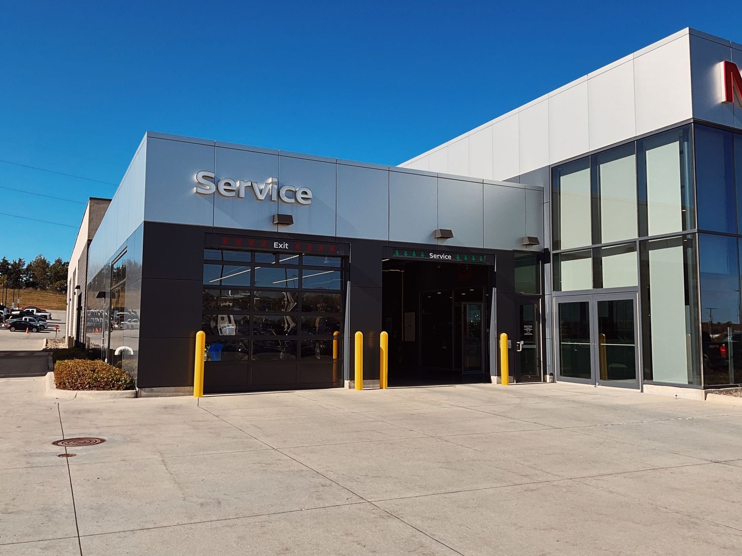 Modern automotive service center exterior with yellow bollards, large glass doors, and Service signage on a sunny day.