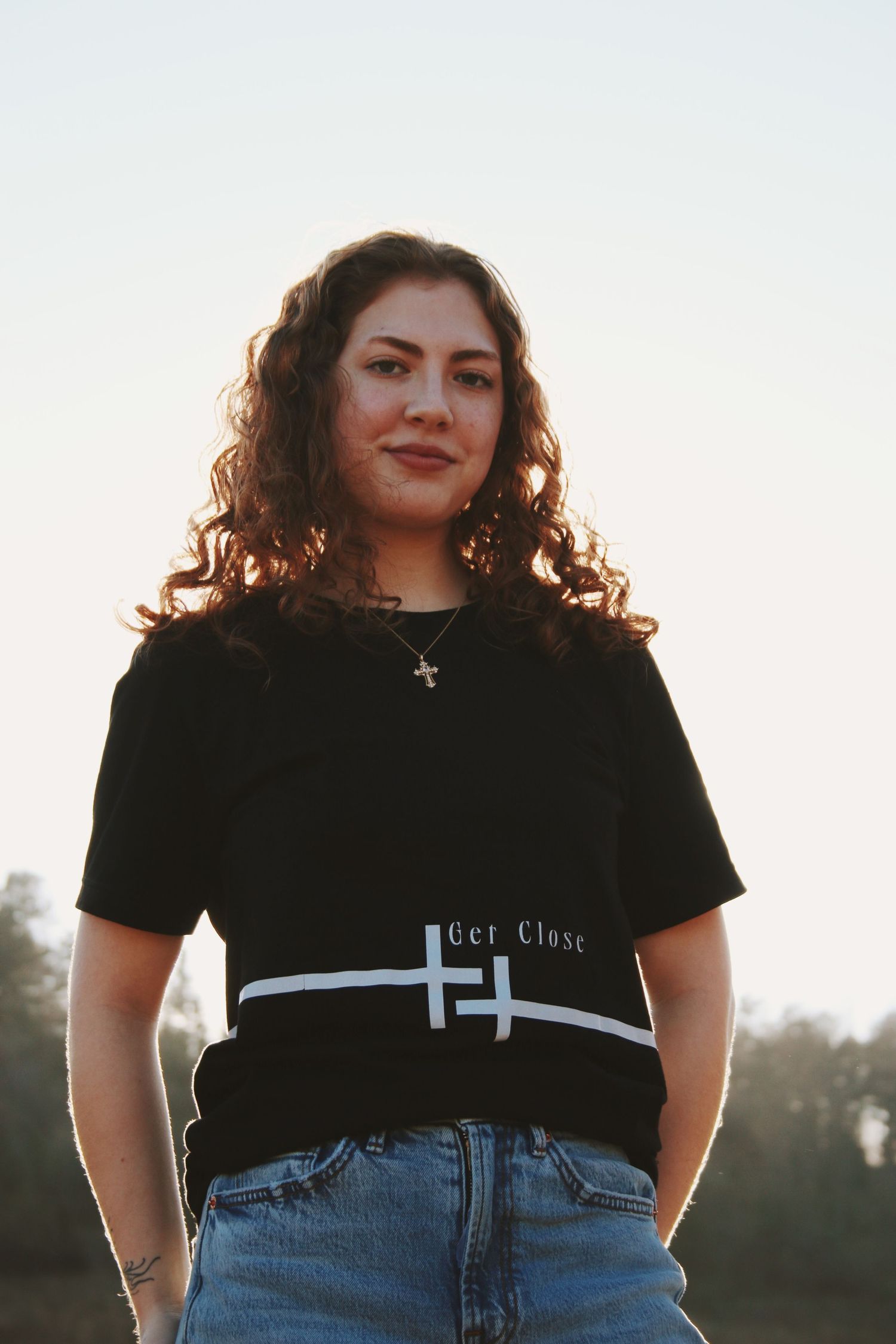 Young woman with curly hair wearing a black graphic tee and jeans, posing outdoors at golden hour.