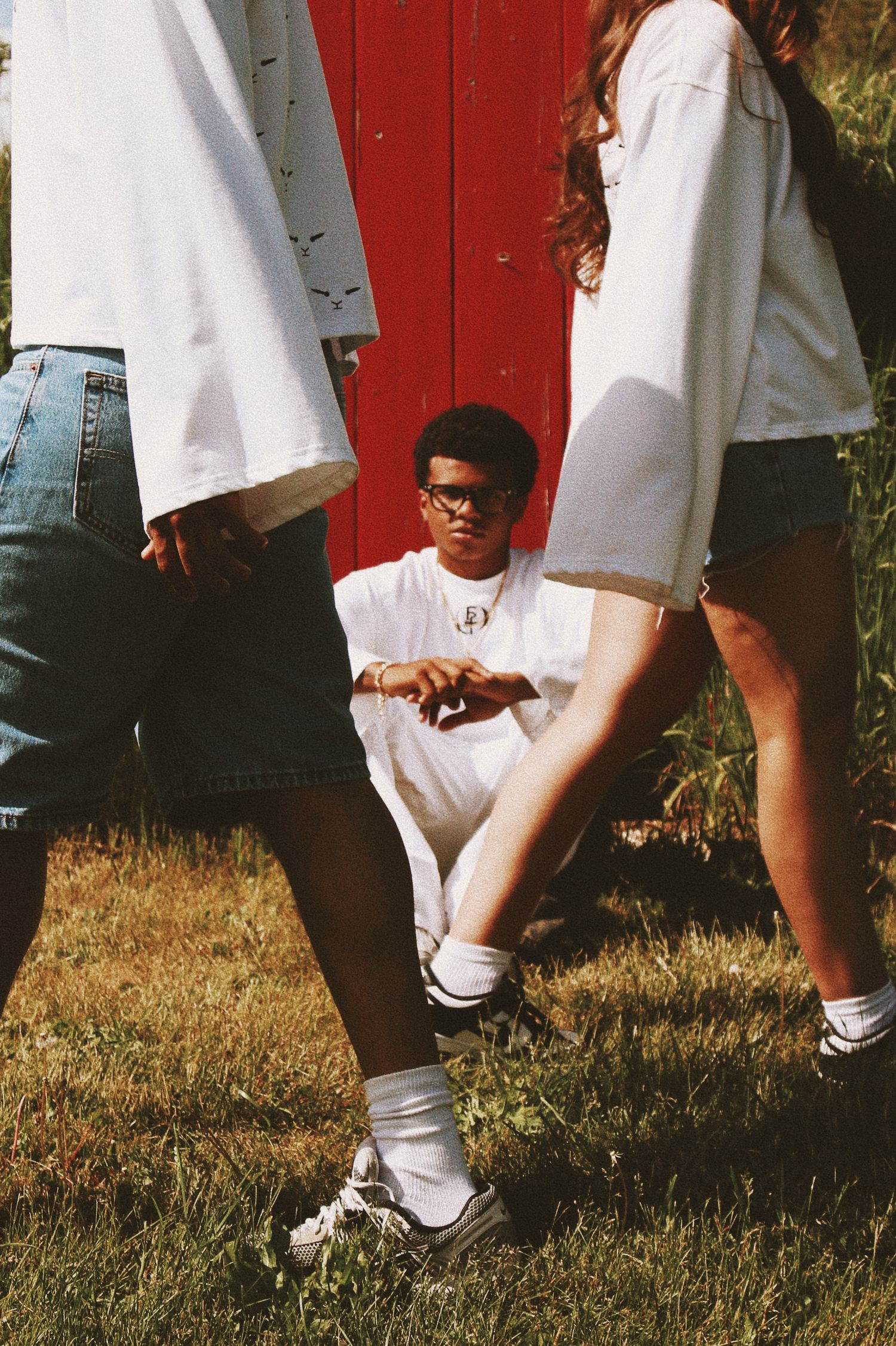 Young man in white tee and glasses crouching outdoors between two people, in front of a red door.