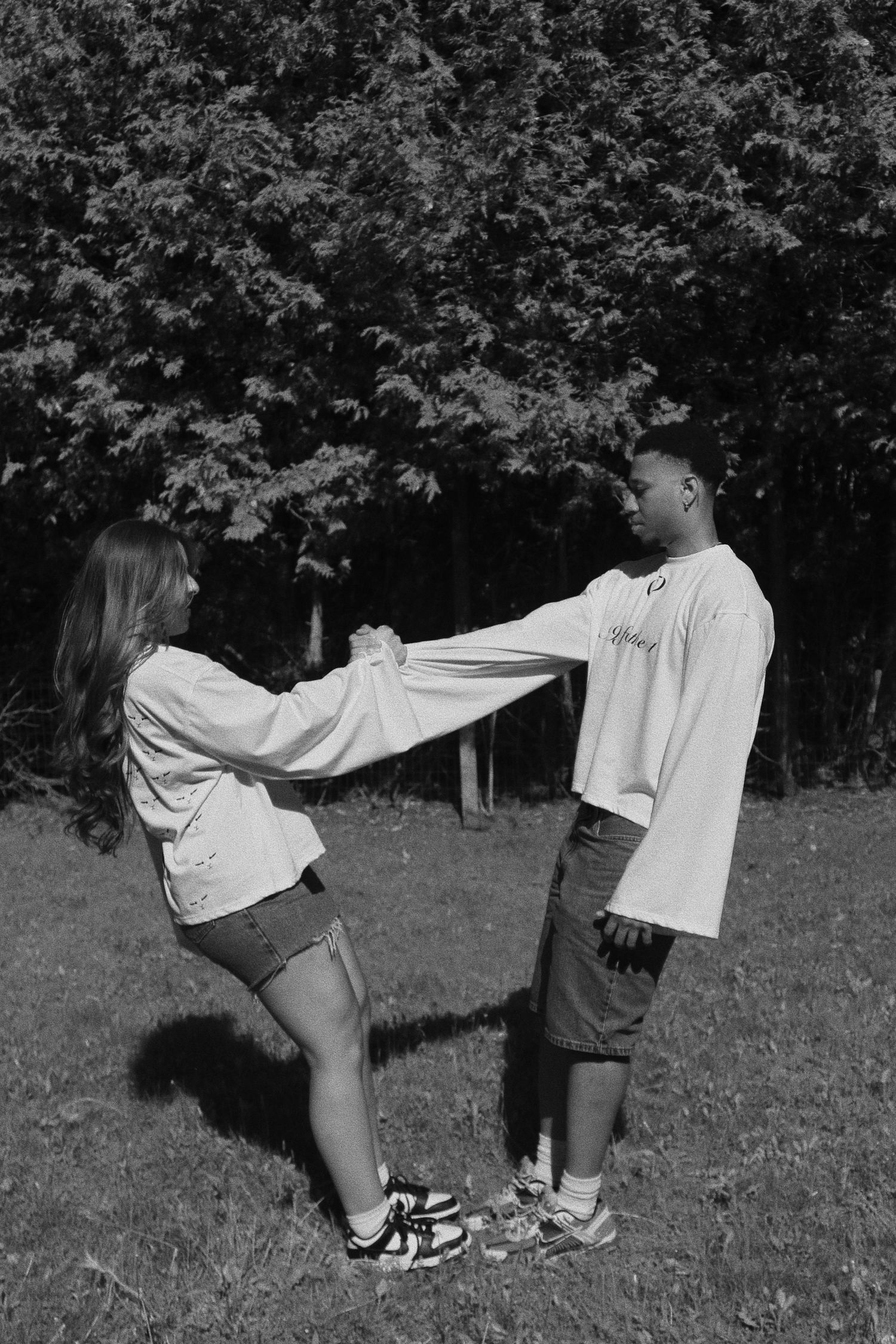 Black and white photo of a couple holding hands and leaning back in a grassy park with trees in the background.