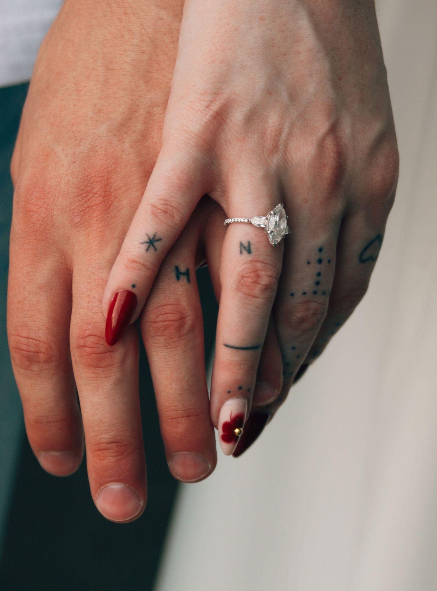 Two hands intertwined showing matching letter tattoos and a diamond engagement ring, with red and neutral manicured nails.