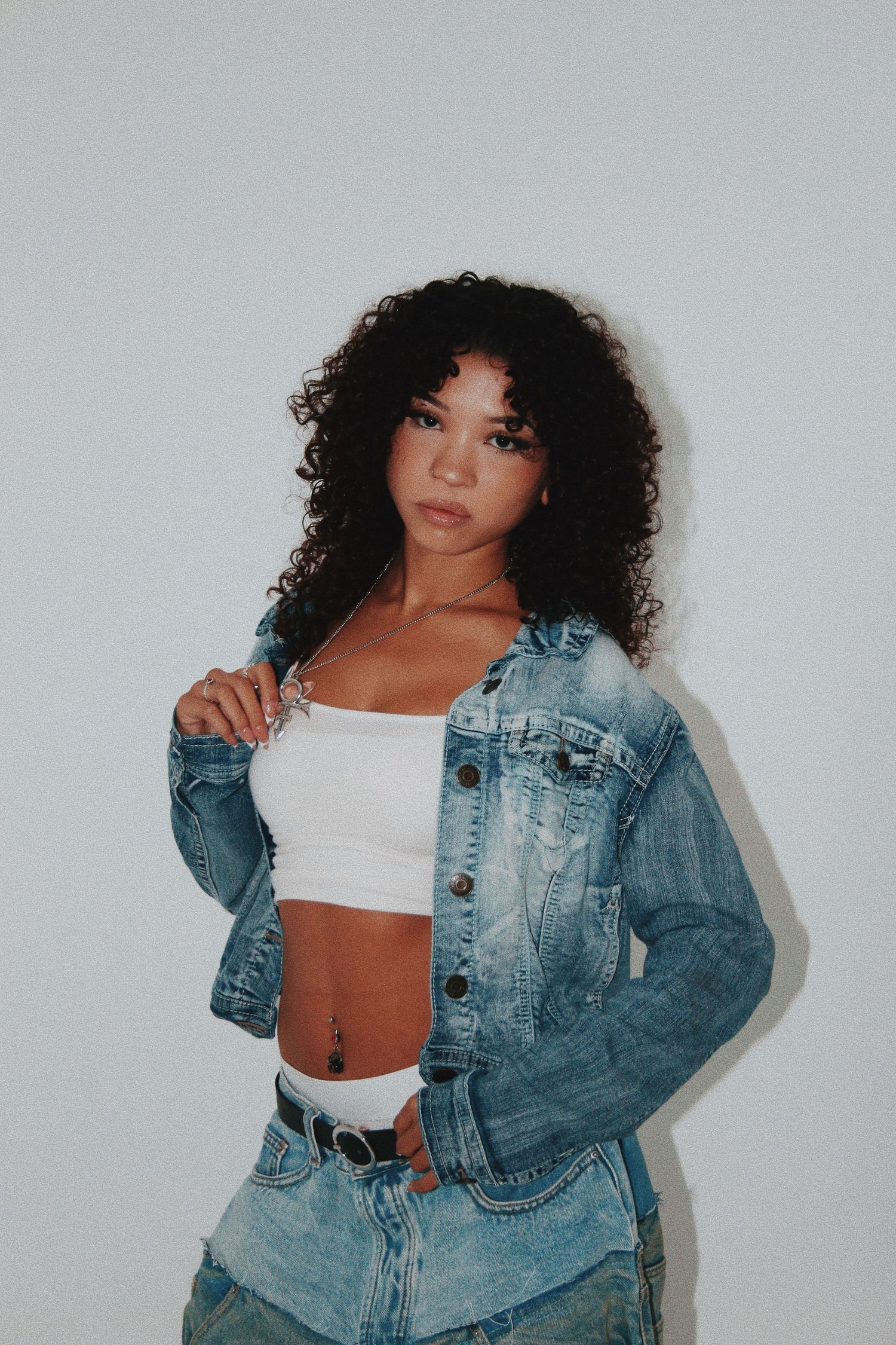 Young woman with curly hair wearing a white crop top and denim jacket poses confidently against a white wall.