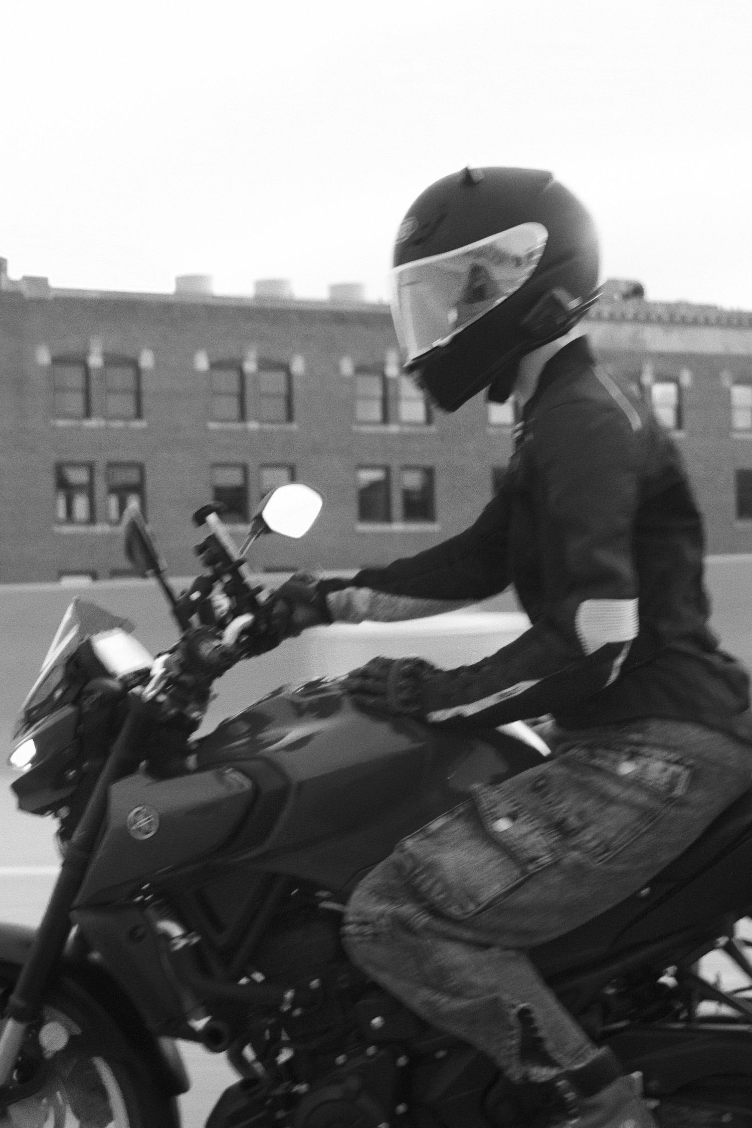 Black and white photo of a motorcyclist wearing a full-face helmet riding a motorcycle past a brick building.