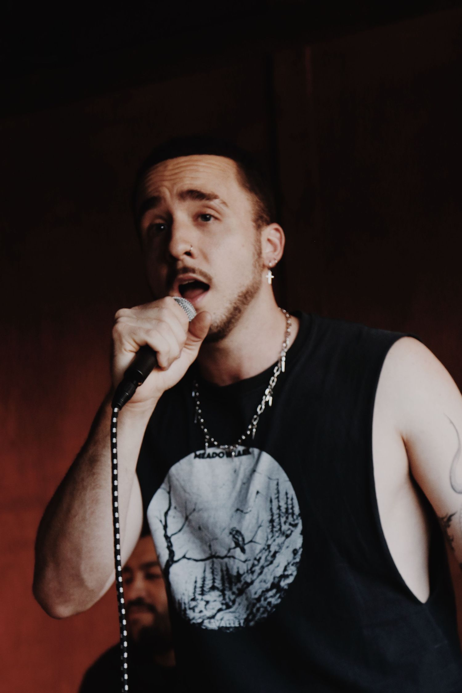Young male vocalist performing on stage, holding microphone, wearing black sleeveless shirt with chain necklace.