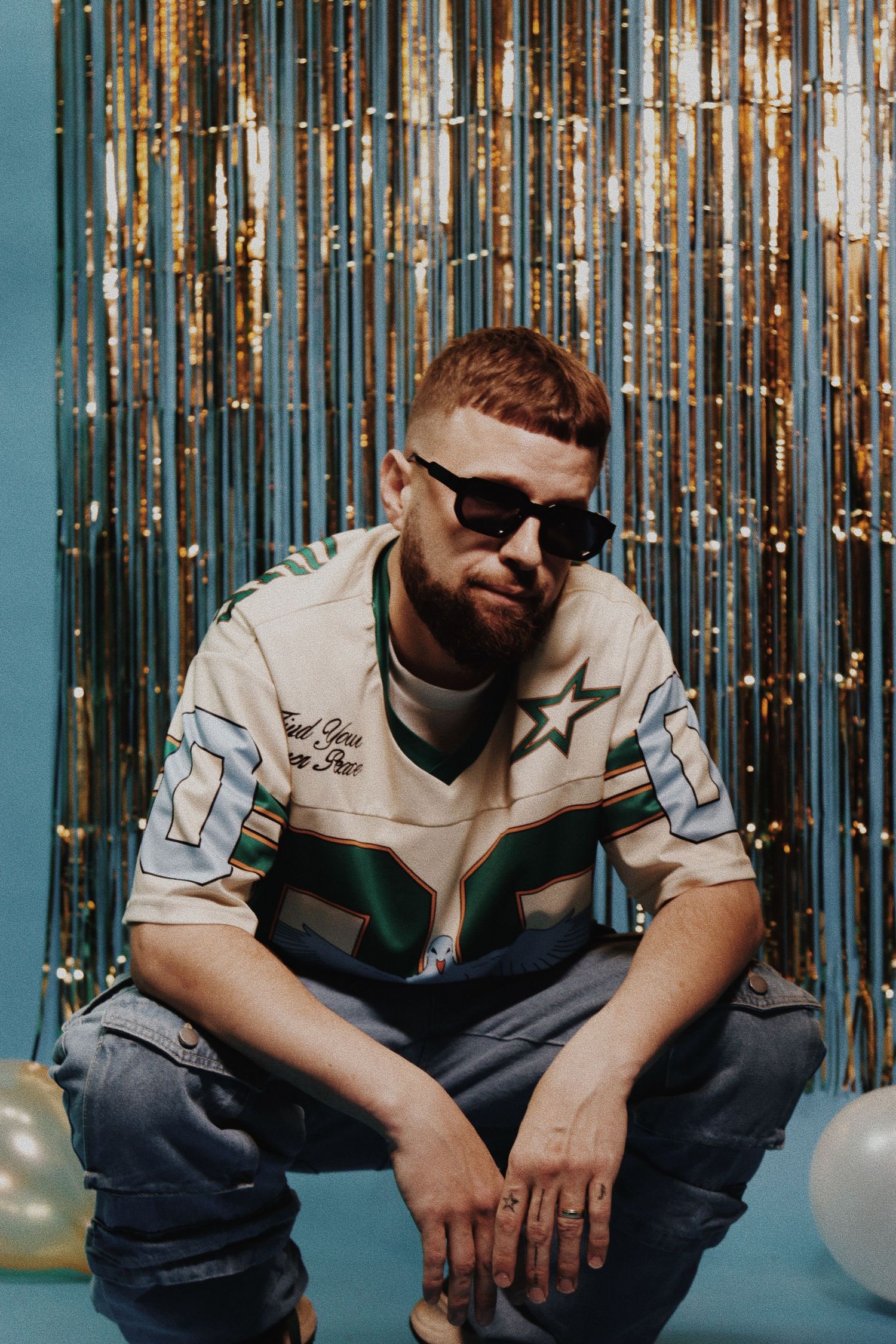 Bearded man in sunglasses and a star-print jersey sits confidently in front of a gold tinsel backdrop.