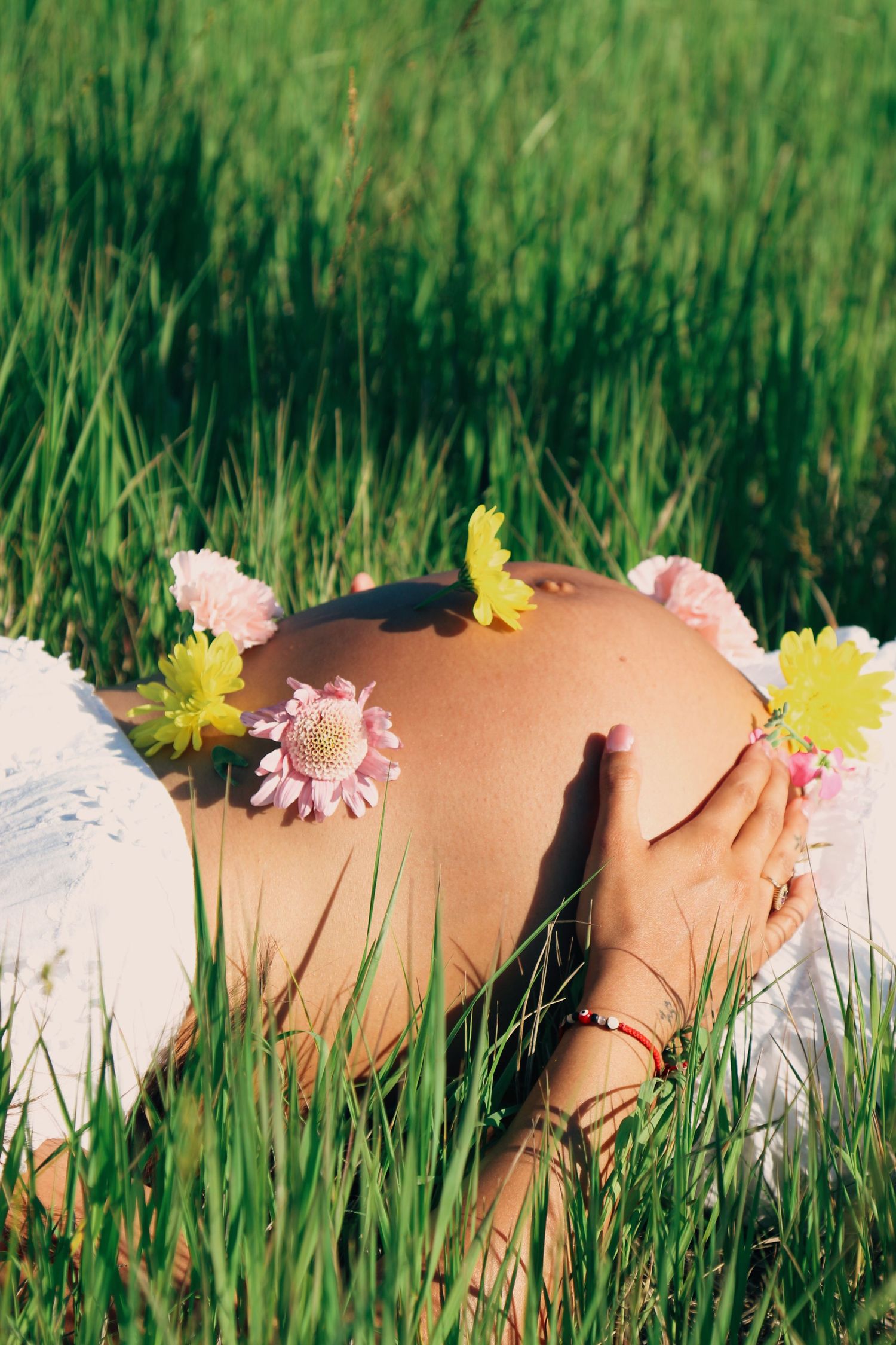 Pregnant woman lying in green grass with colorful daisy flowers resting on her bare baby bump on a sunny day.