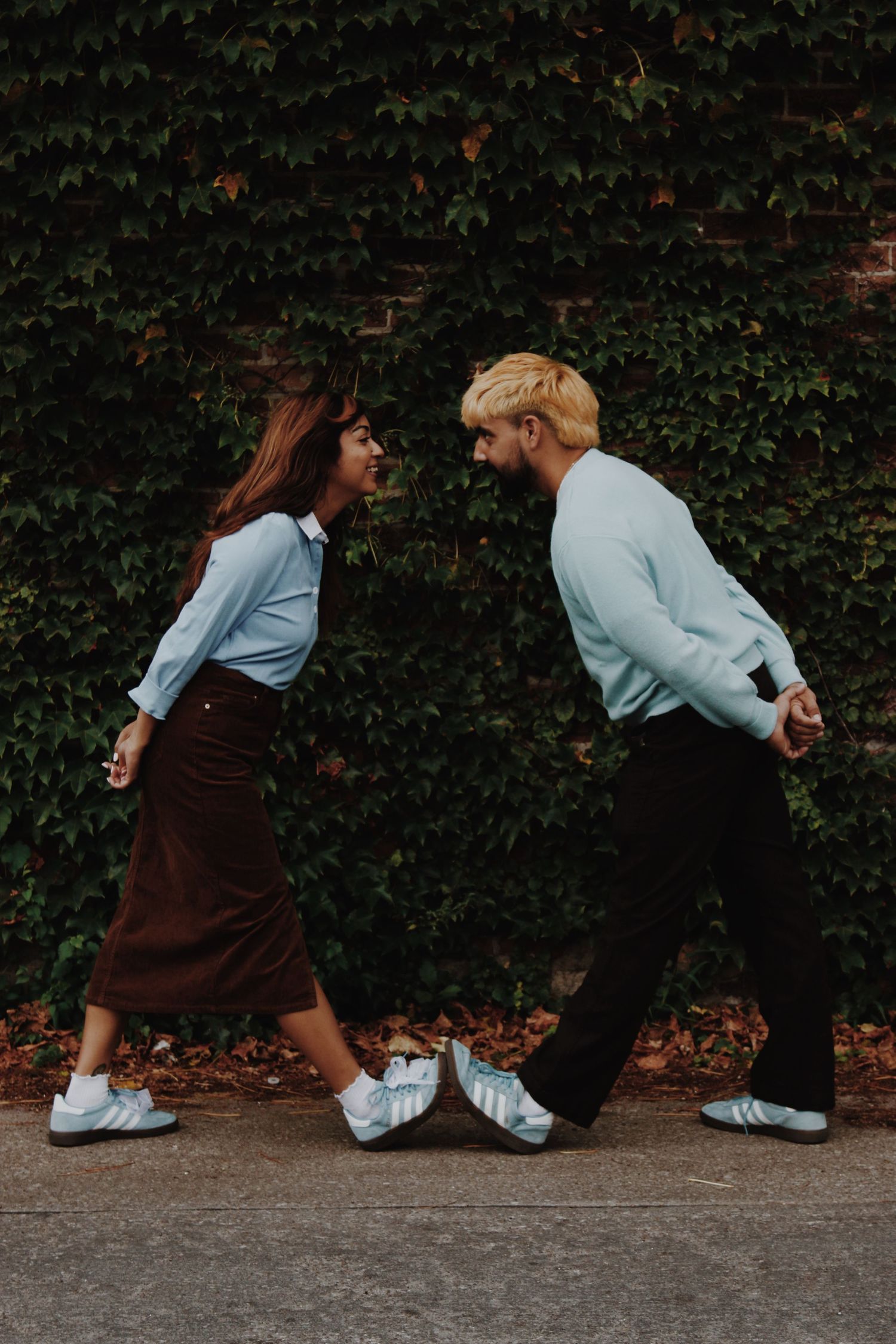 A young couple in matching light blue tops and white Adidas sneakers face each other smiling against an ivy-covered brick wall.