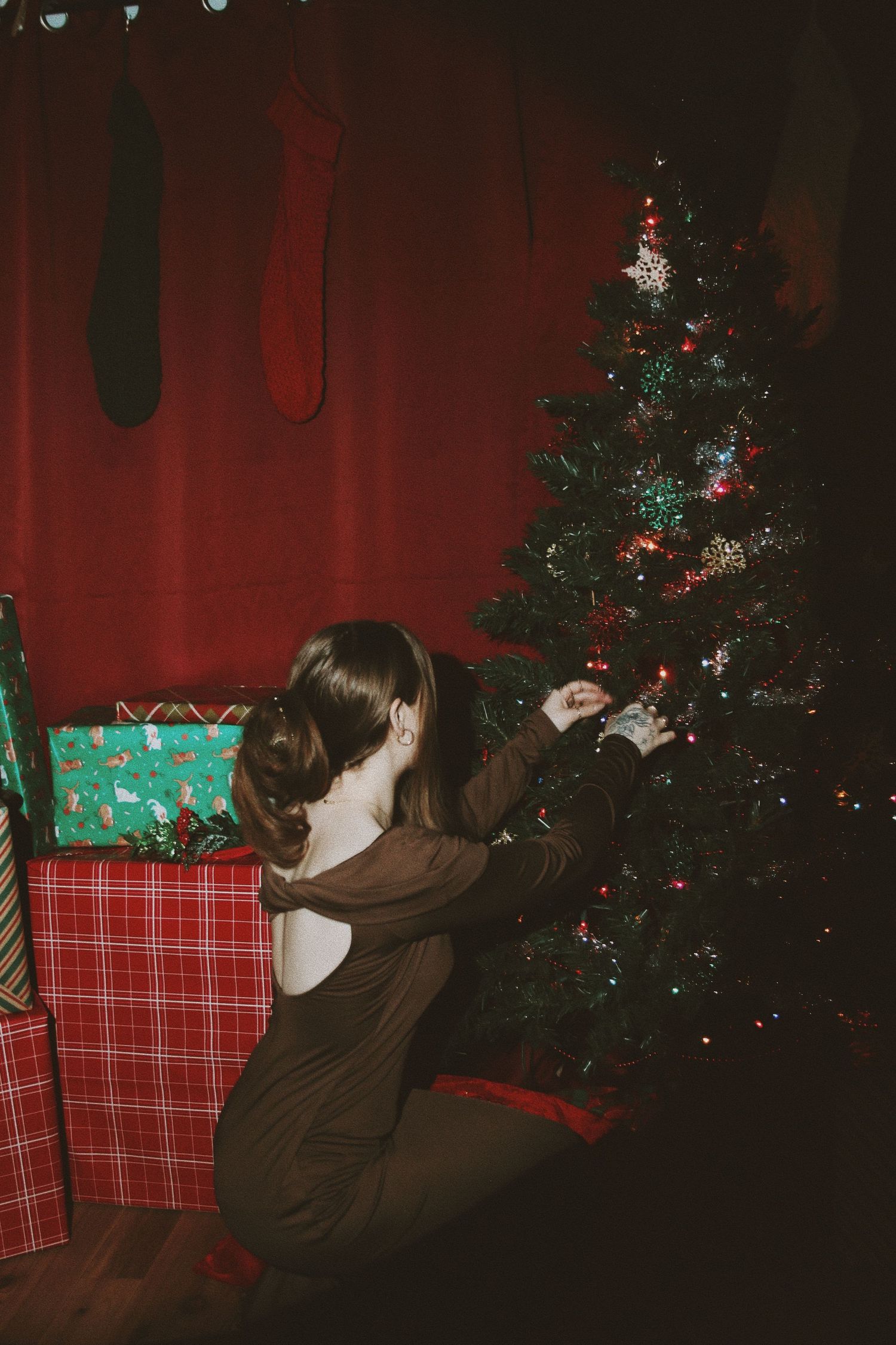 Young woman in a brown dress decorating a Christmas tree surrounded by wrapped gifts and holiday stockings.