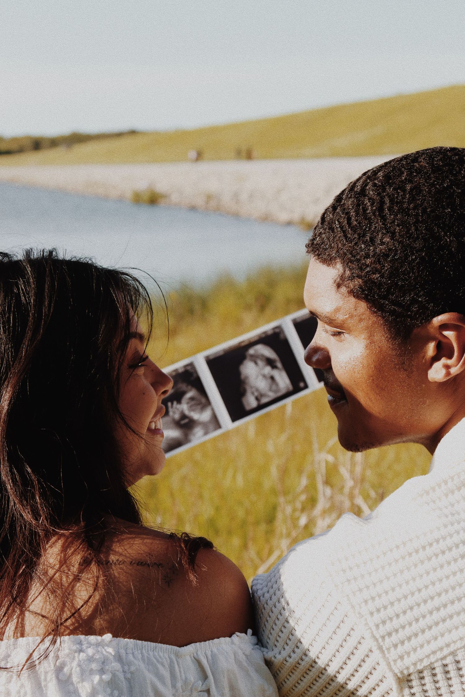 Happy couple holding ultrasound photos together near a scenic lakeside meadow on a sunny day.
