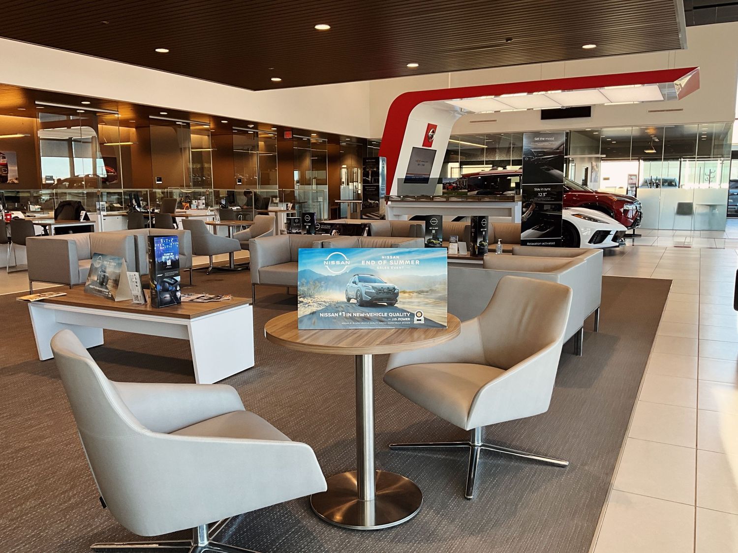 Modern car dealership showroom with white chairs, tablet display table, and luxury vehicles on the floor.