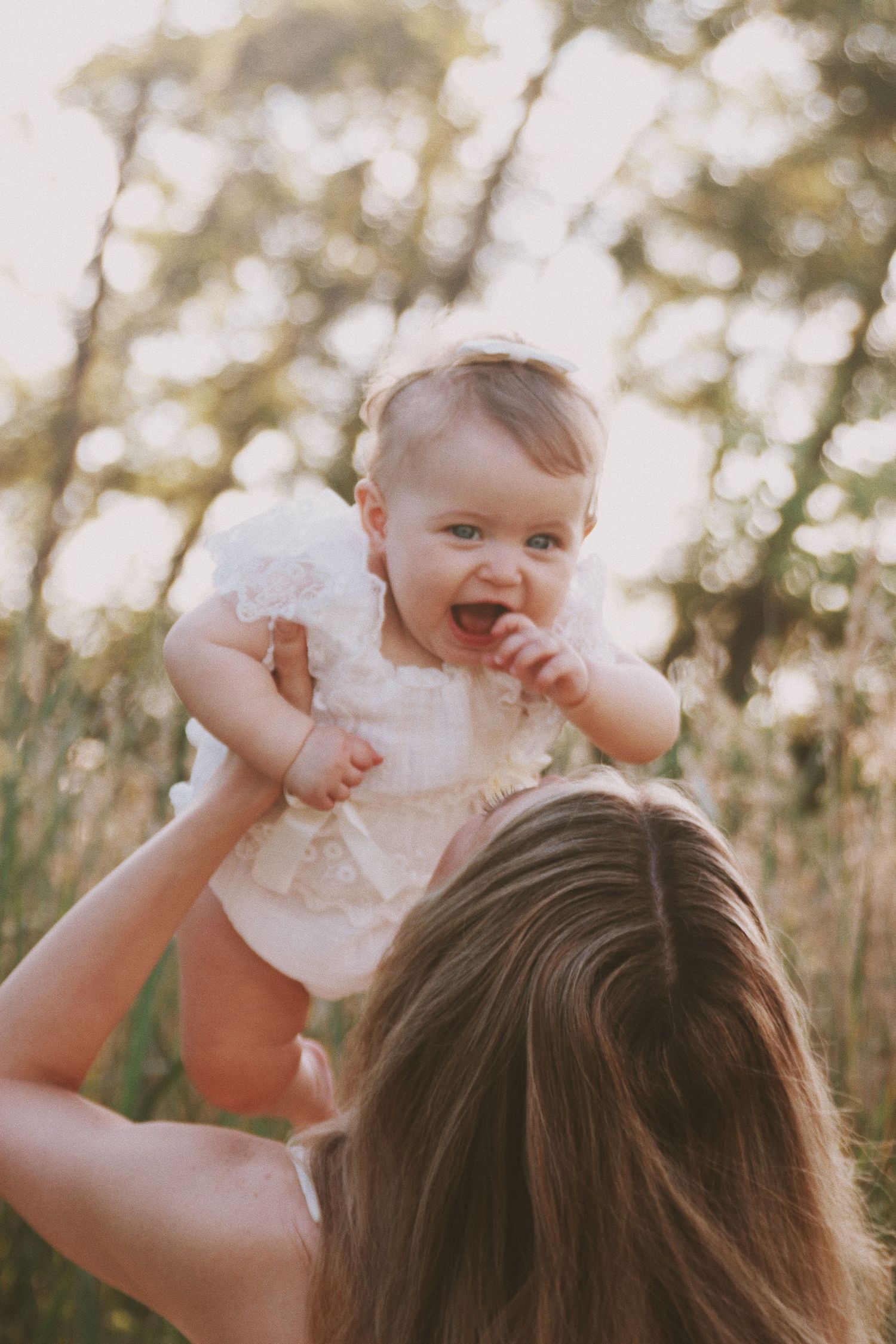 A joyful baby in a white lace dress being lifted by a mother outdoors in a sunlit green field.