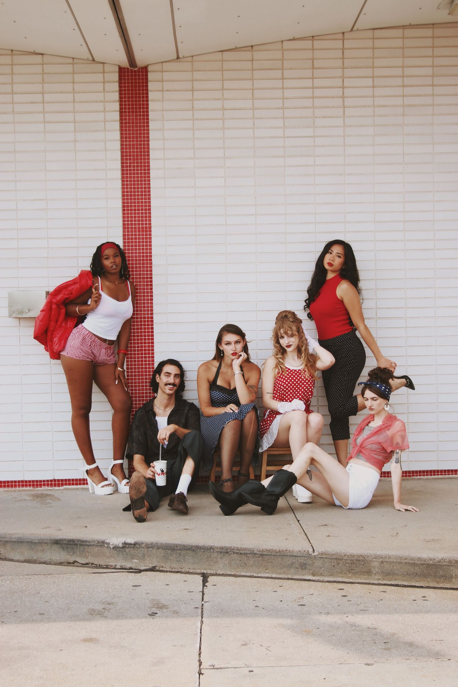 Six stylish young adults pose in retro-inspired outfits outside a diner with red and white tile decor.