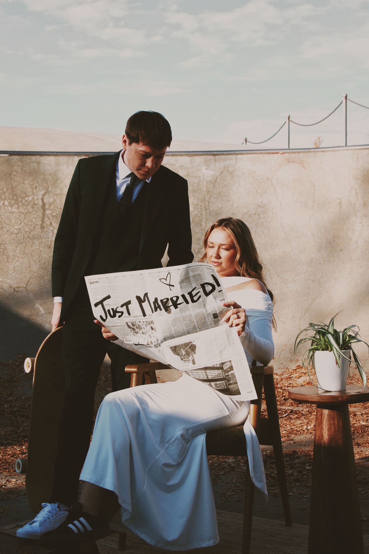 Newlywed couple holding a Just Married sign outdoors, bride in white dress seated while groom stands beside her.