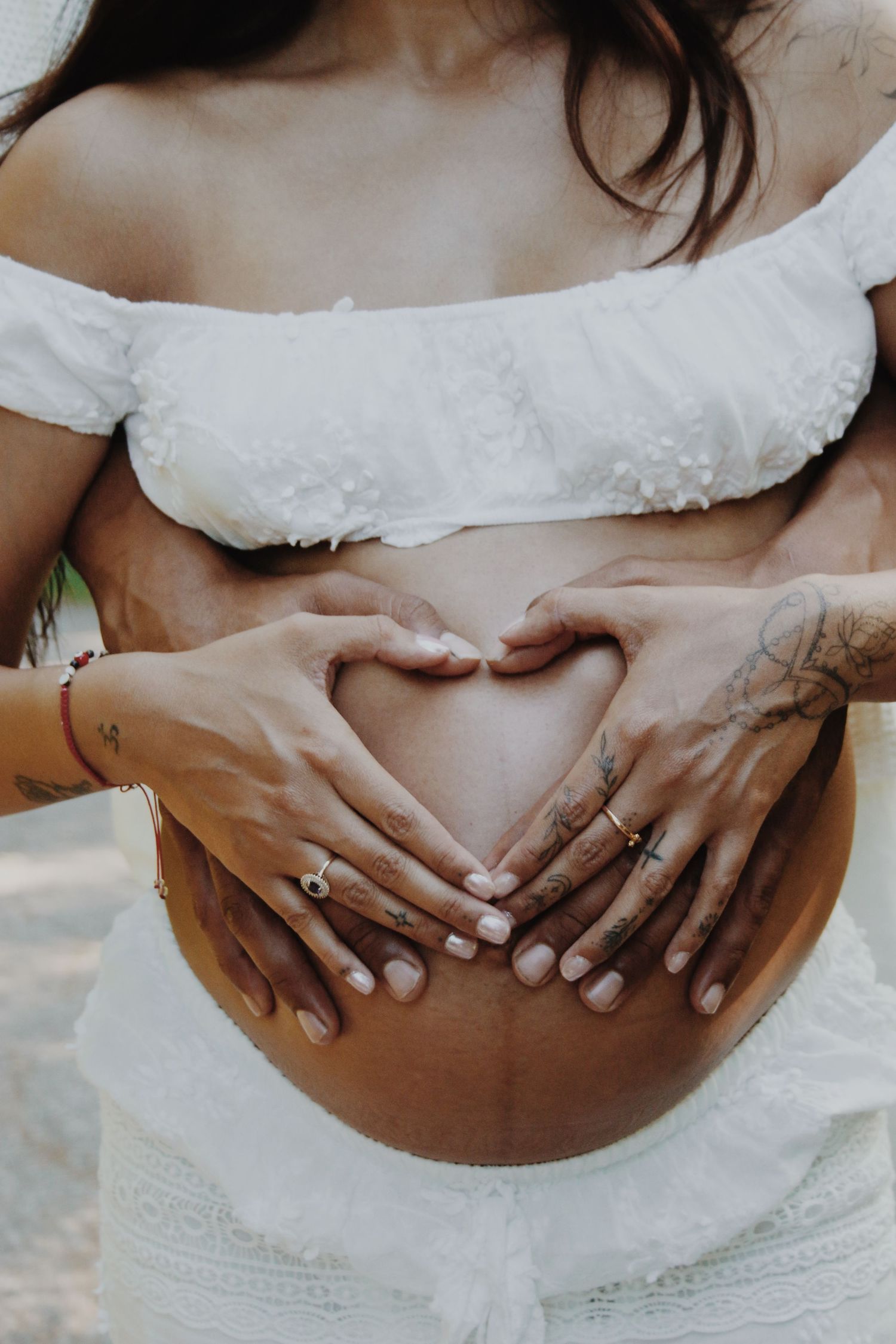 Two hands forming a heart shape on a pregnant woman's bare belly, maternity photo shoot.