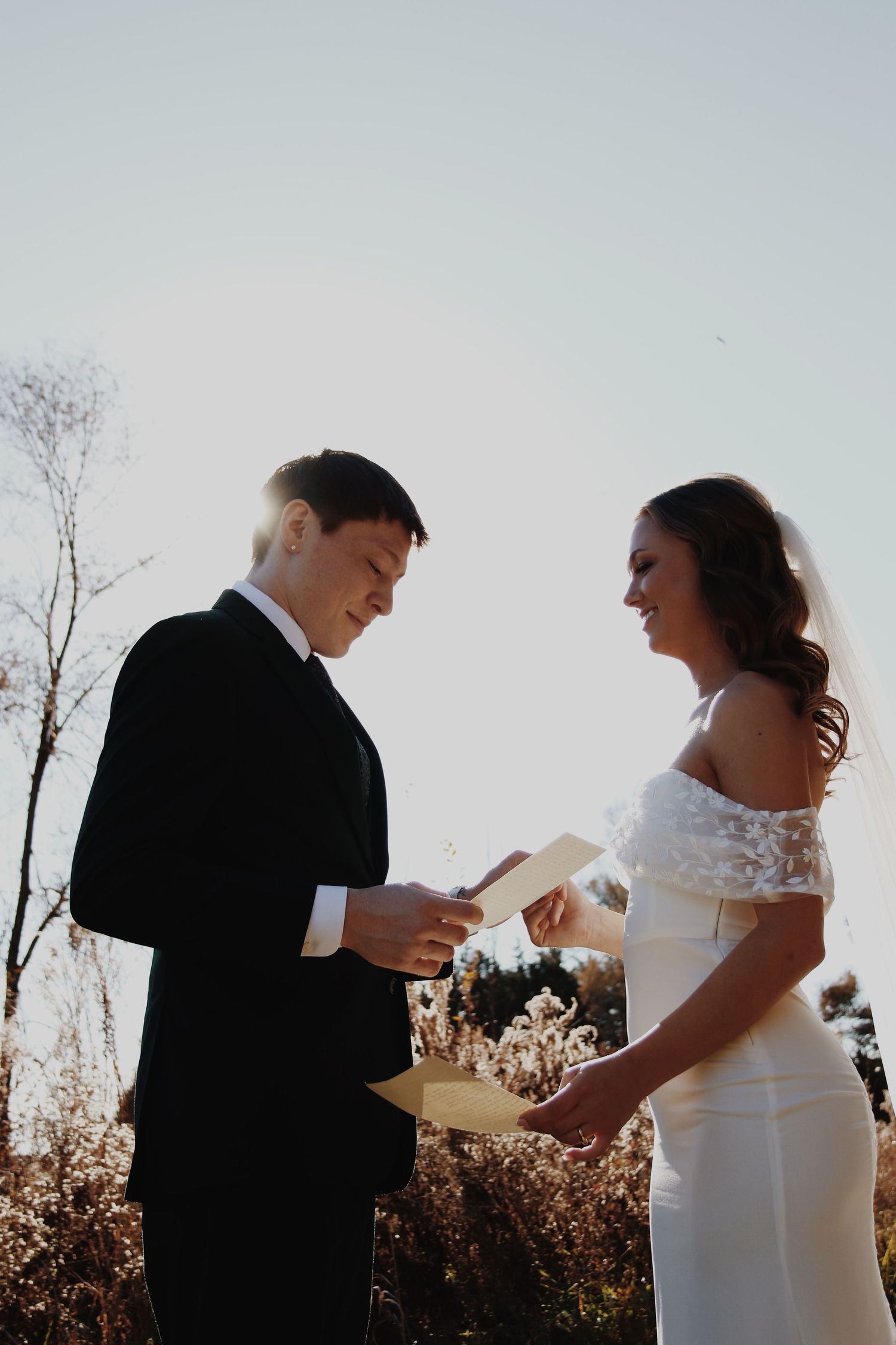 Bride and groom exchange vows outdoors, reading from cards during a sunlit wedding ceremony.