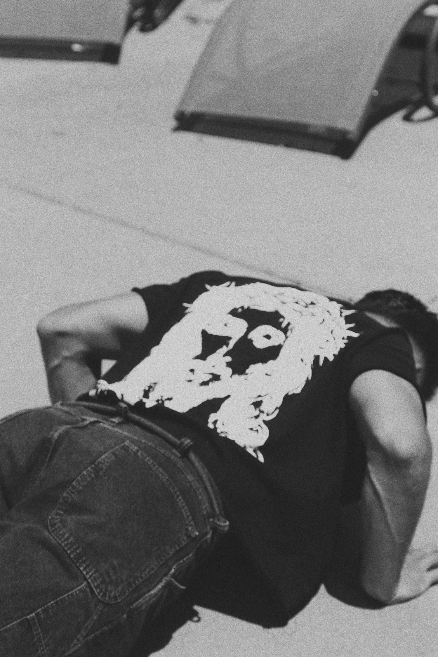 Black and white photo of a person lying face down on concrete wearing a black graphic tee with a face print and jeans.