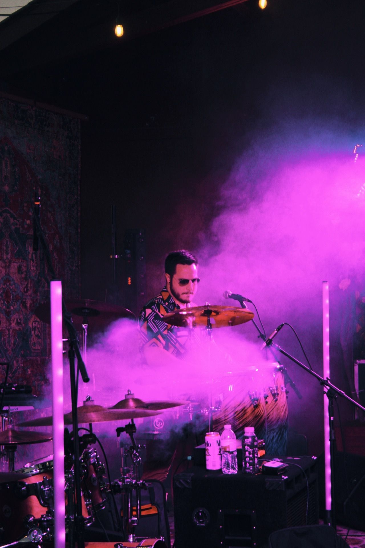 A drummer performs on stage surrounded by dramatic pink smoke and purple lighting during a live music concert.