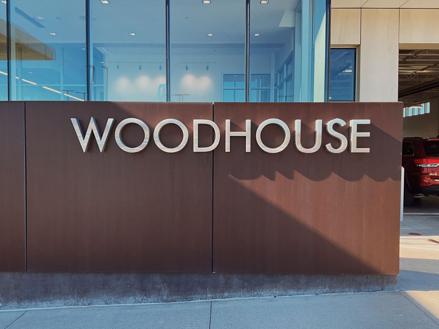 Woodhouse dealership exterior sign mounted on a modern brown metal wall with glass building facade in the background.