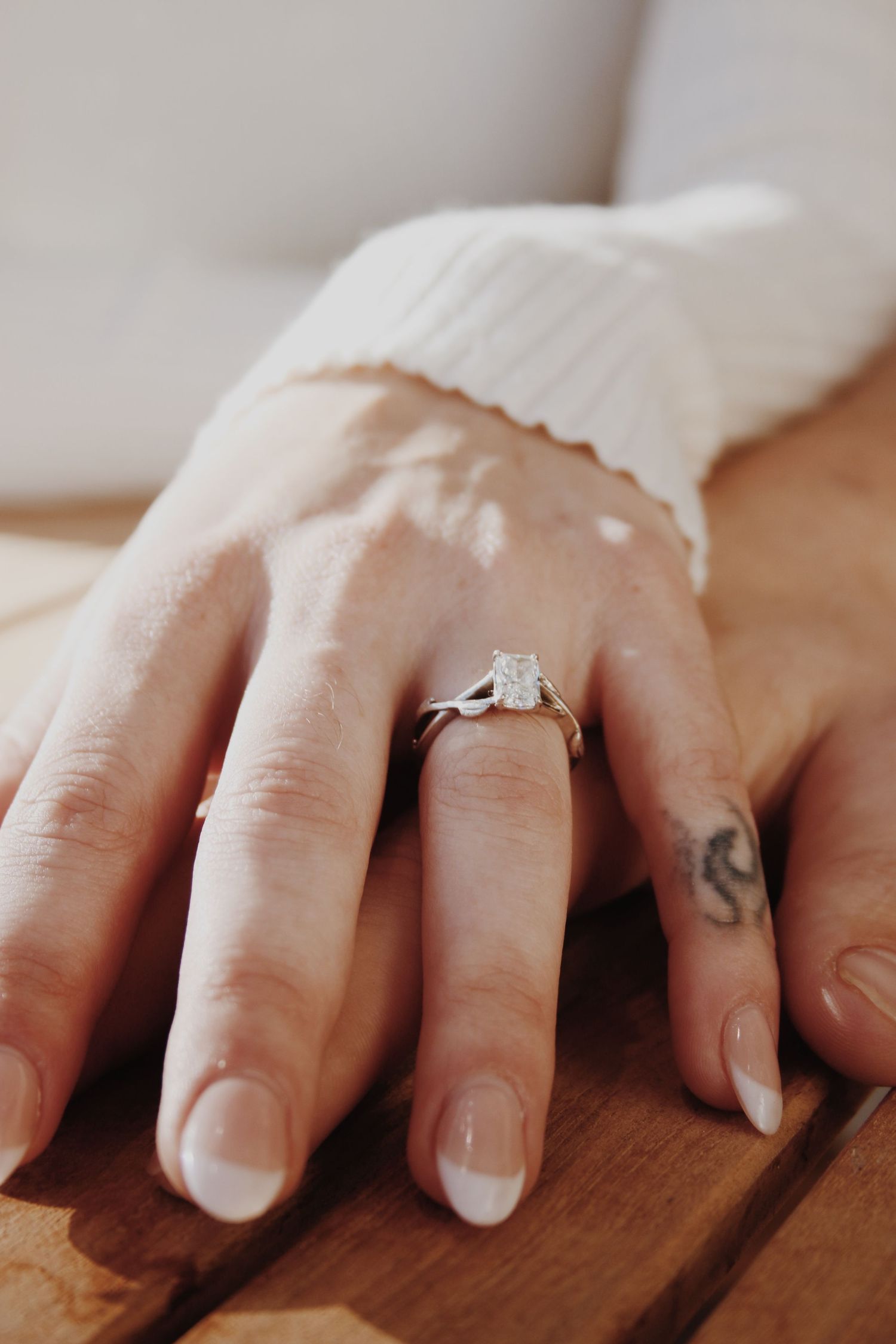 Close-up of a hand wearing a solitaire diamond engagement ring on ring finger, with small tattoo visible.