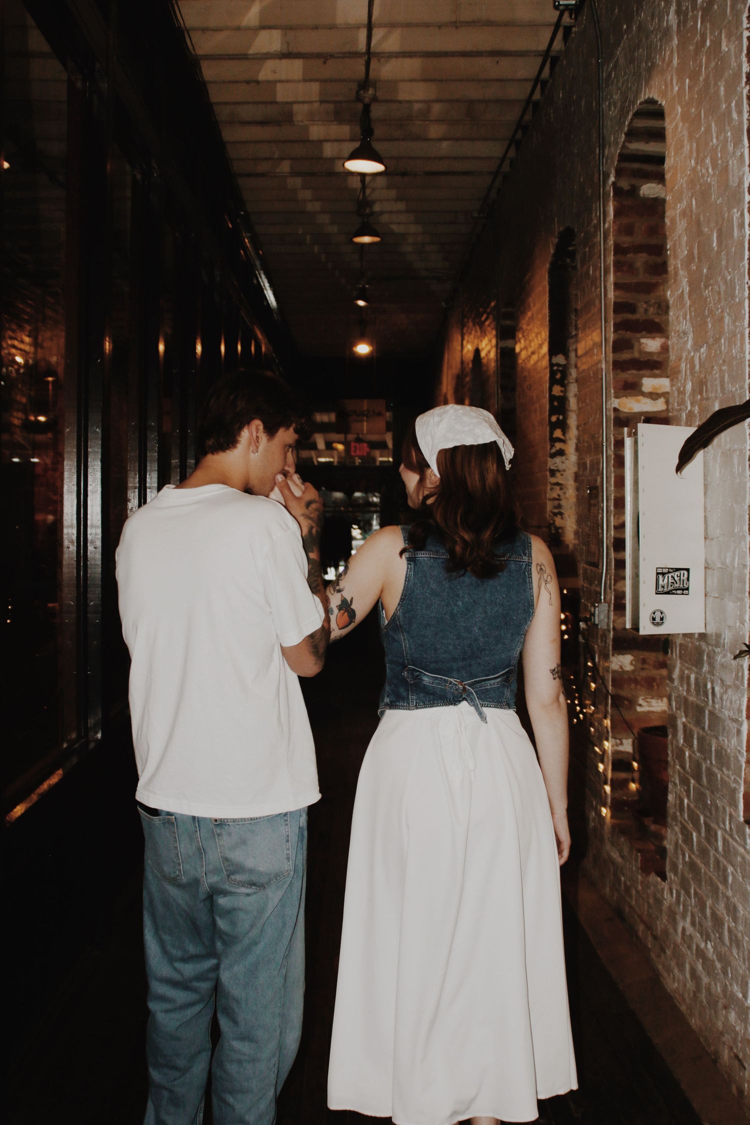 Two people standing in a dimly lit brick corridor, woman wearing white skirt and cap with denim top.