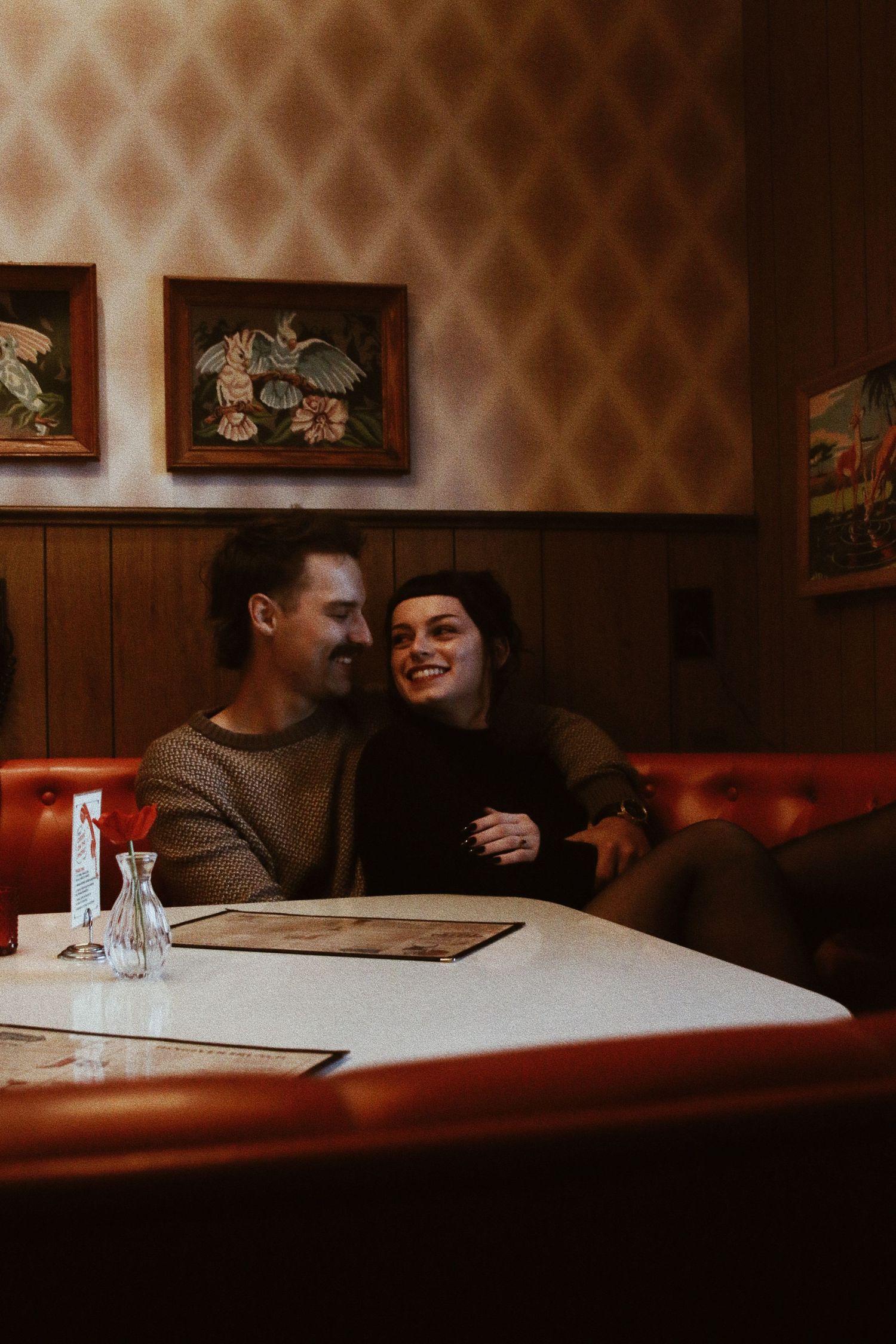 Couple smiling and laughing together in a cozy retro diner booth with vintage wallpaper and framed artwork.