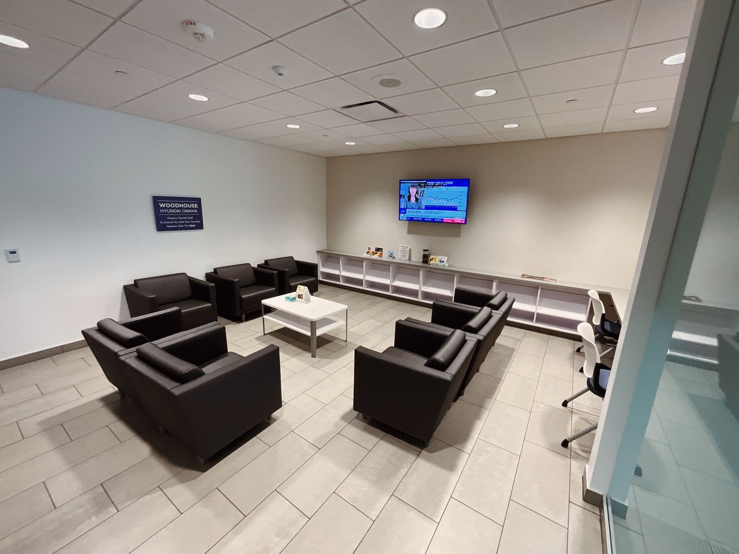 Modern office waiting room with black sofas, white coffee table, wall-mounted TV, and shelving unit on tiled floor.