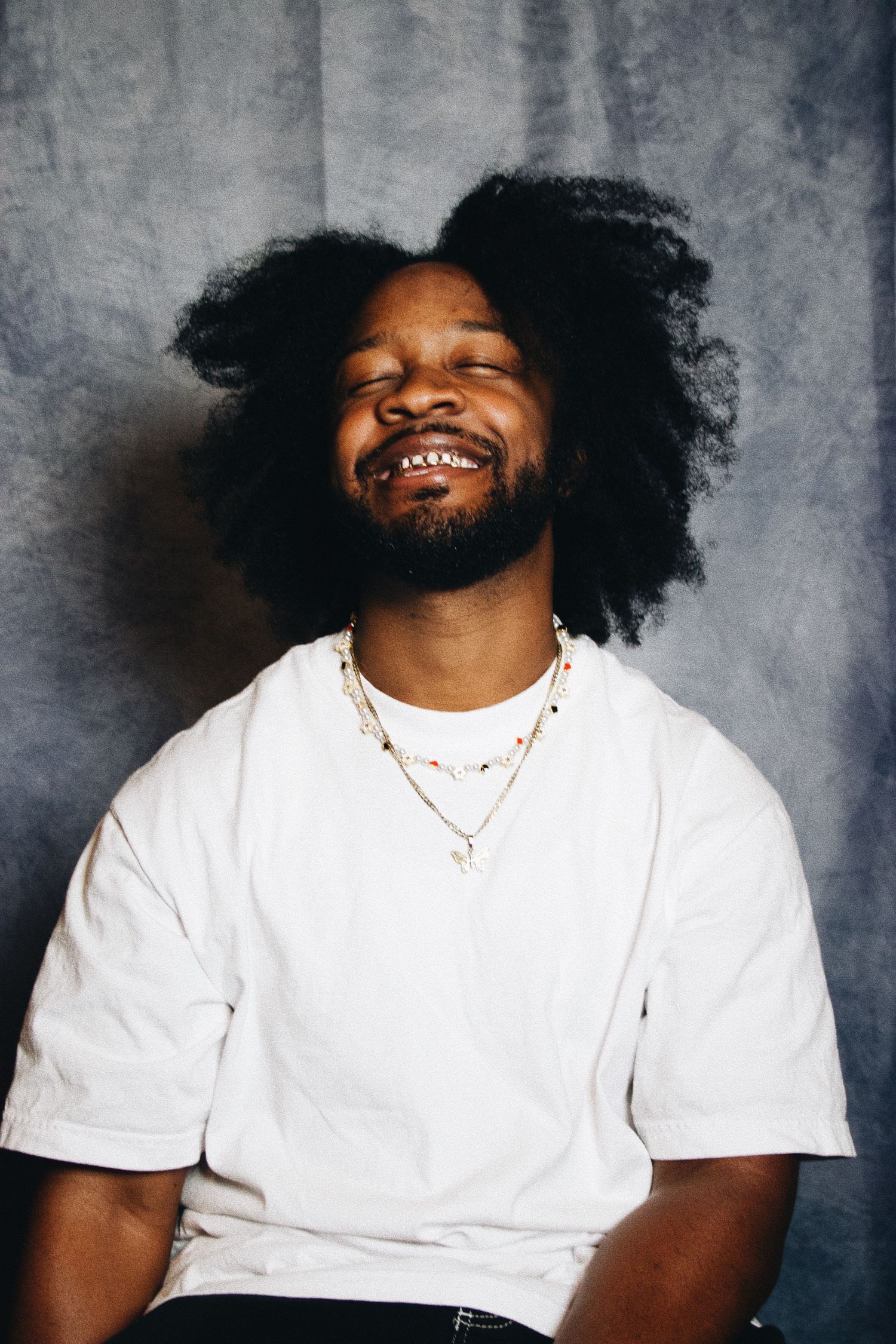 Joyful Black man with afro hair, beard, white t-shirt, and layered necklaces laughing with eyes closed.