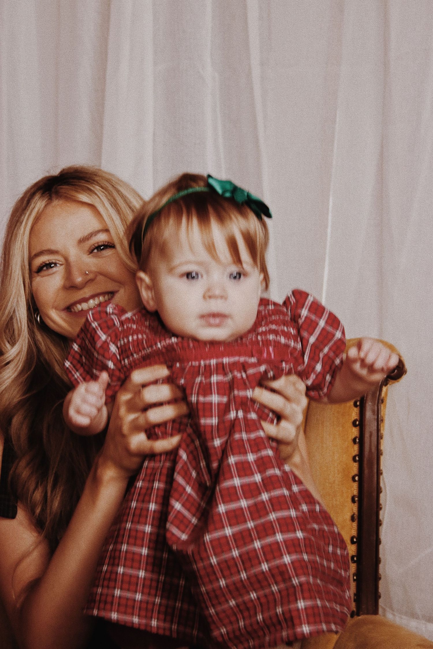 Smiling blonde woman holding a baby girl in a red plaid dress with a green bow on a vintage chair.