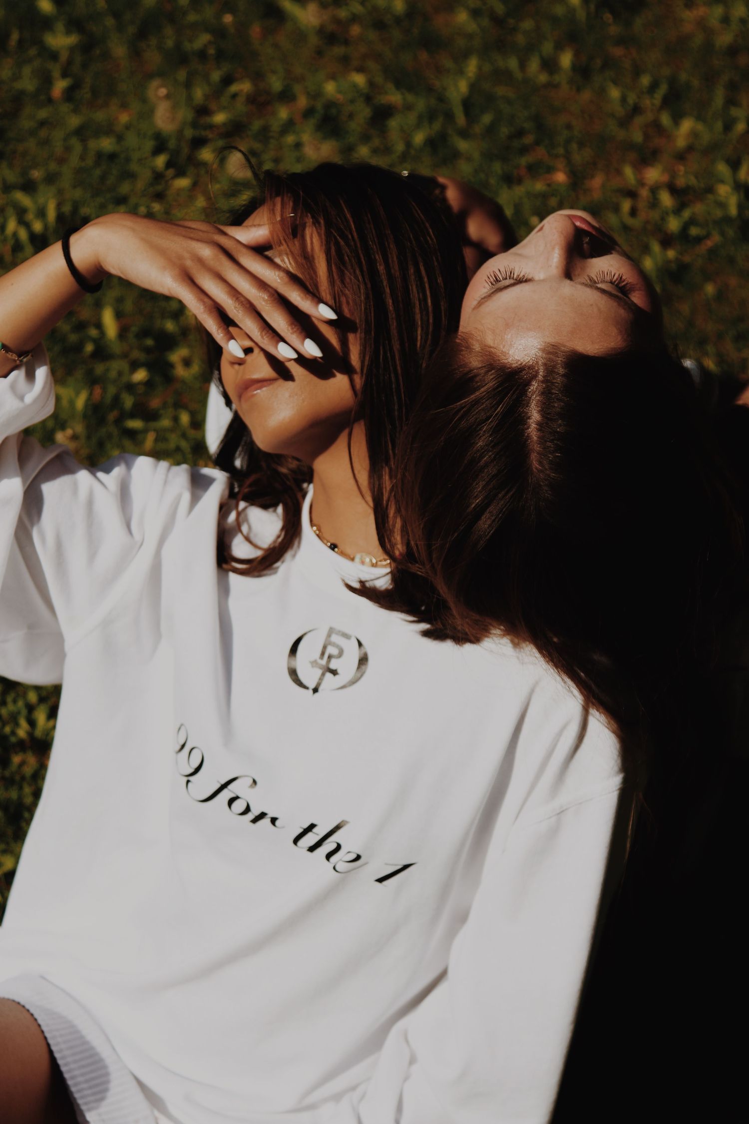 Woman in white graphic long sleeve shirt relaxing outdoors in sunlight, shielding her eyes with her hand.