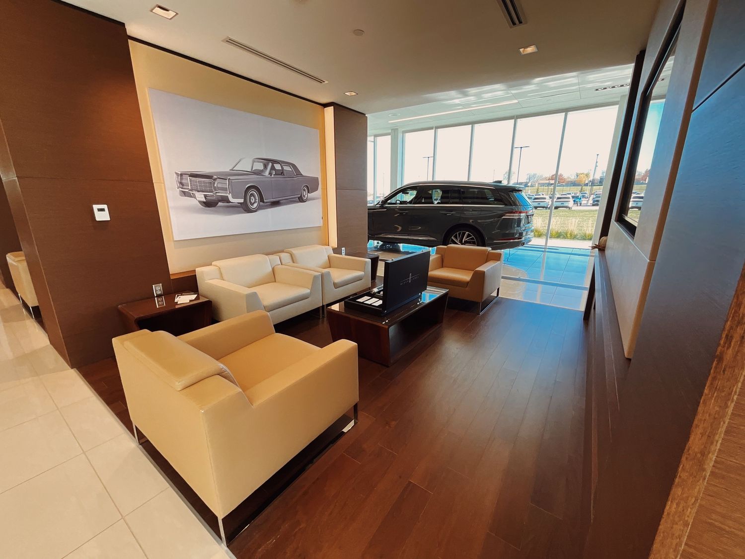 Luxury car dealership showroom with cream sofas, vintage car artwork, and a dark SUV displayed by floor-to-ceiling windows.