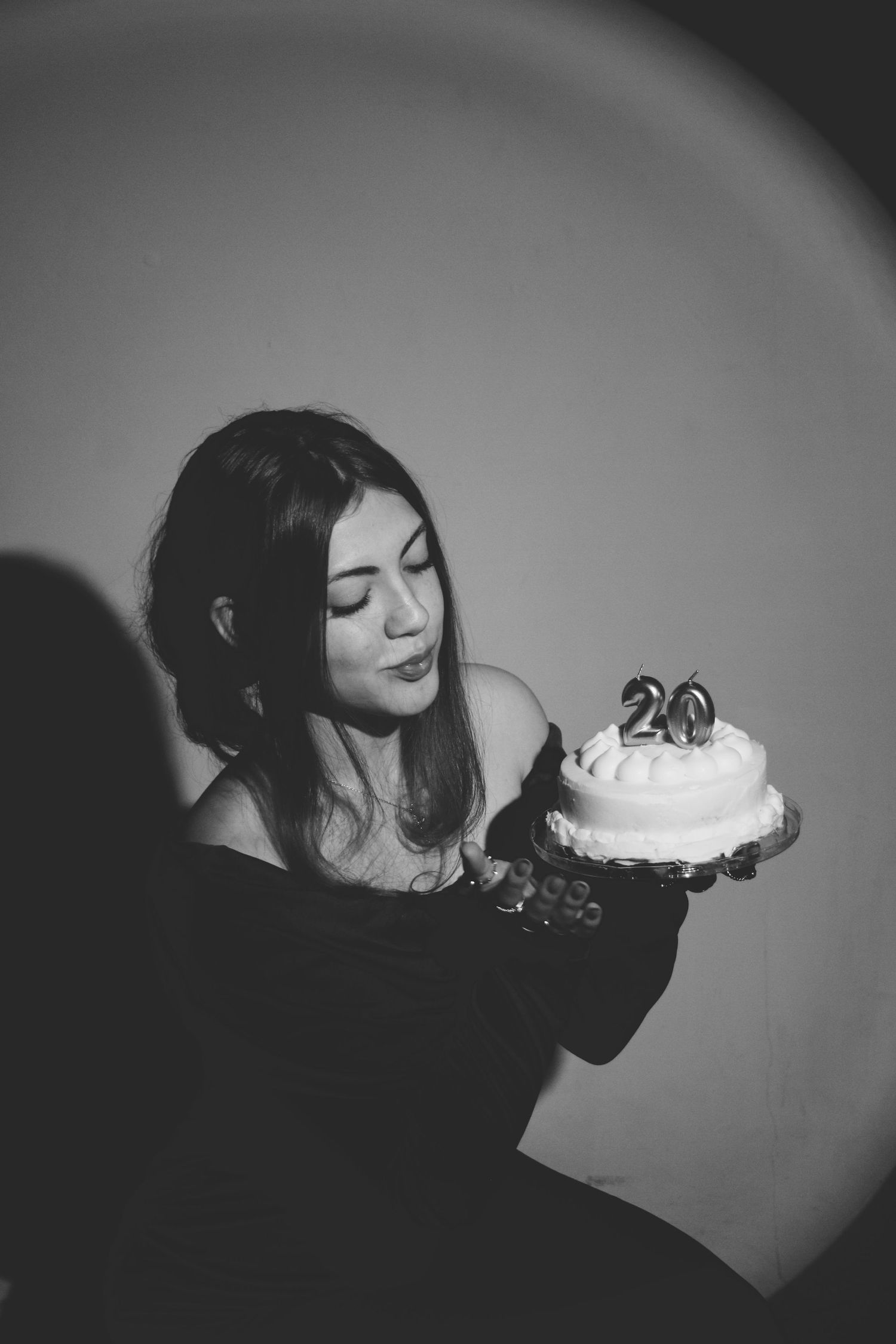 Young woman in black dress blowing out candles on a white birthday cake with number 20, black and white photo.