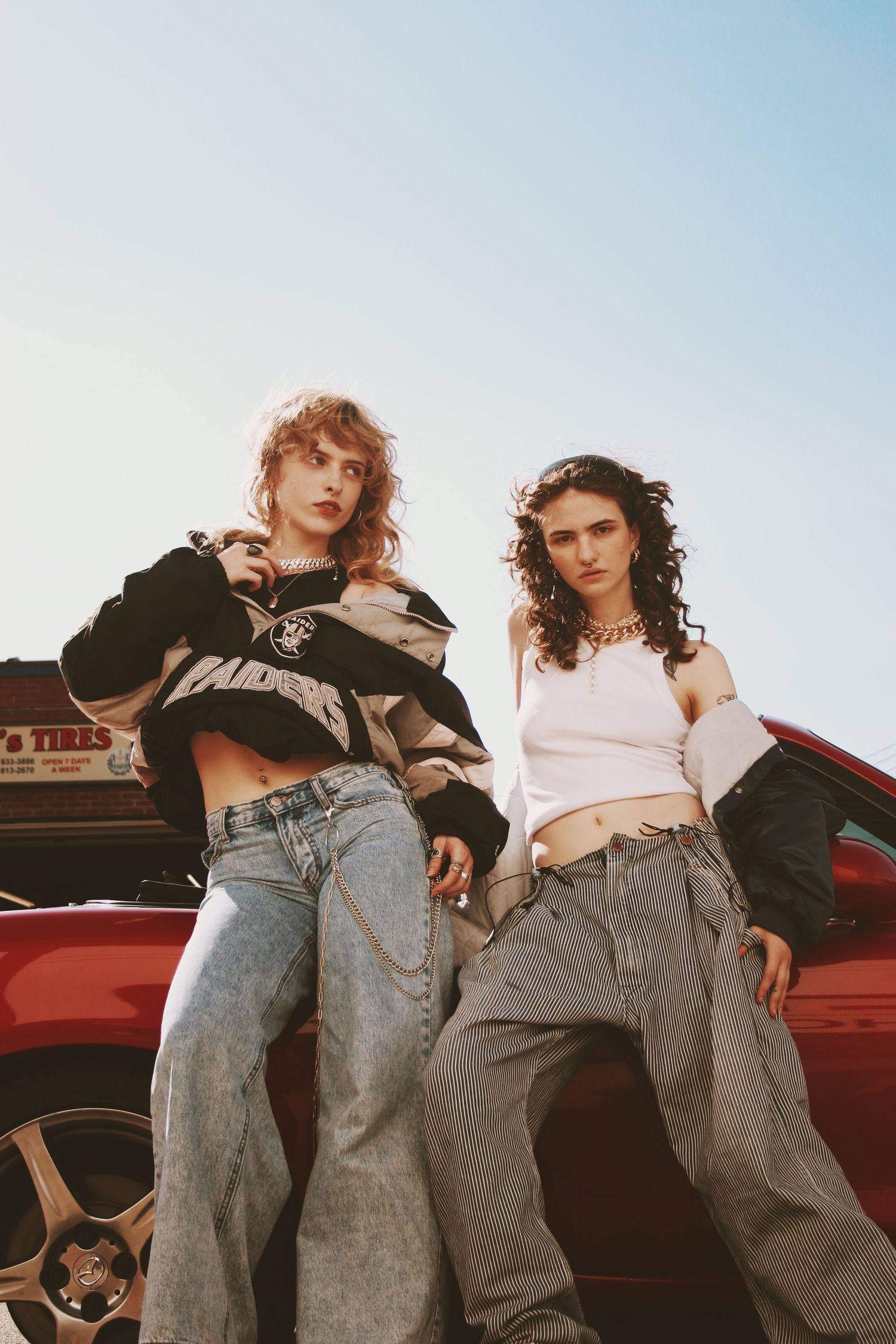 Two women in baggy jeans and crop tops lean against a red car outdoors in a retro 90s-inspired fashion editorial.
