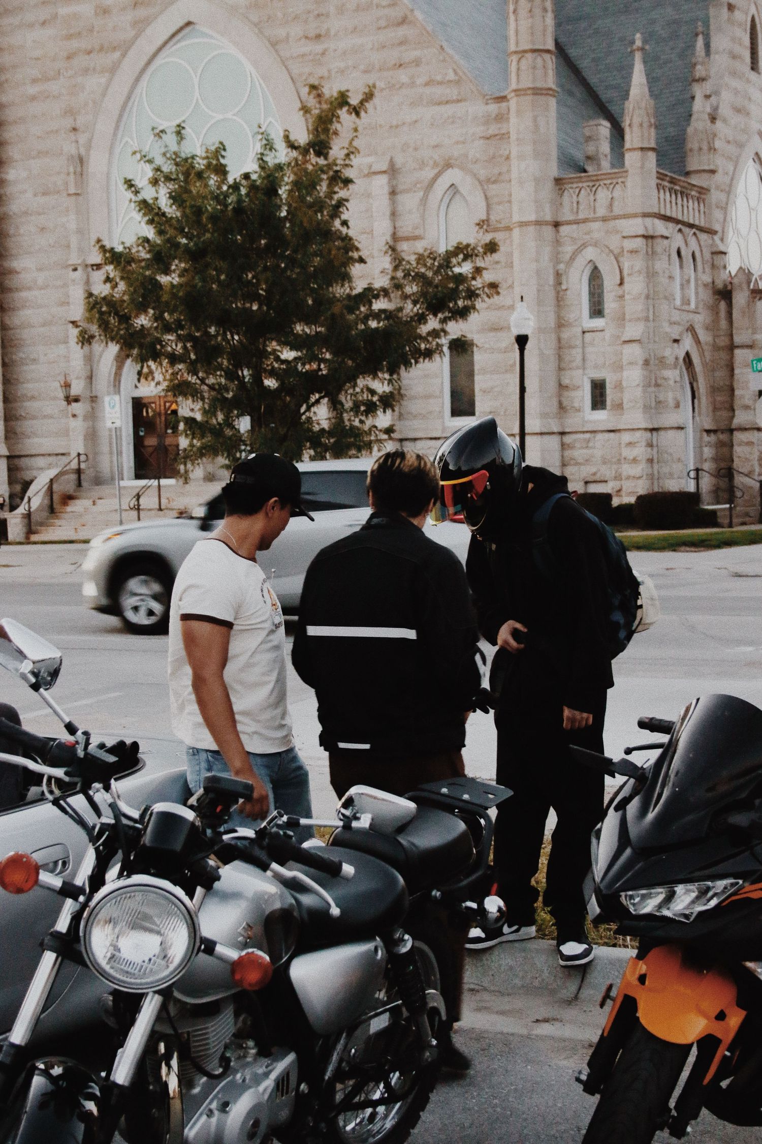 Three motorcyclists gathered near parked bikes outside a historic stone church on a sunny day.