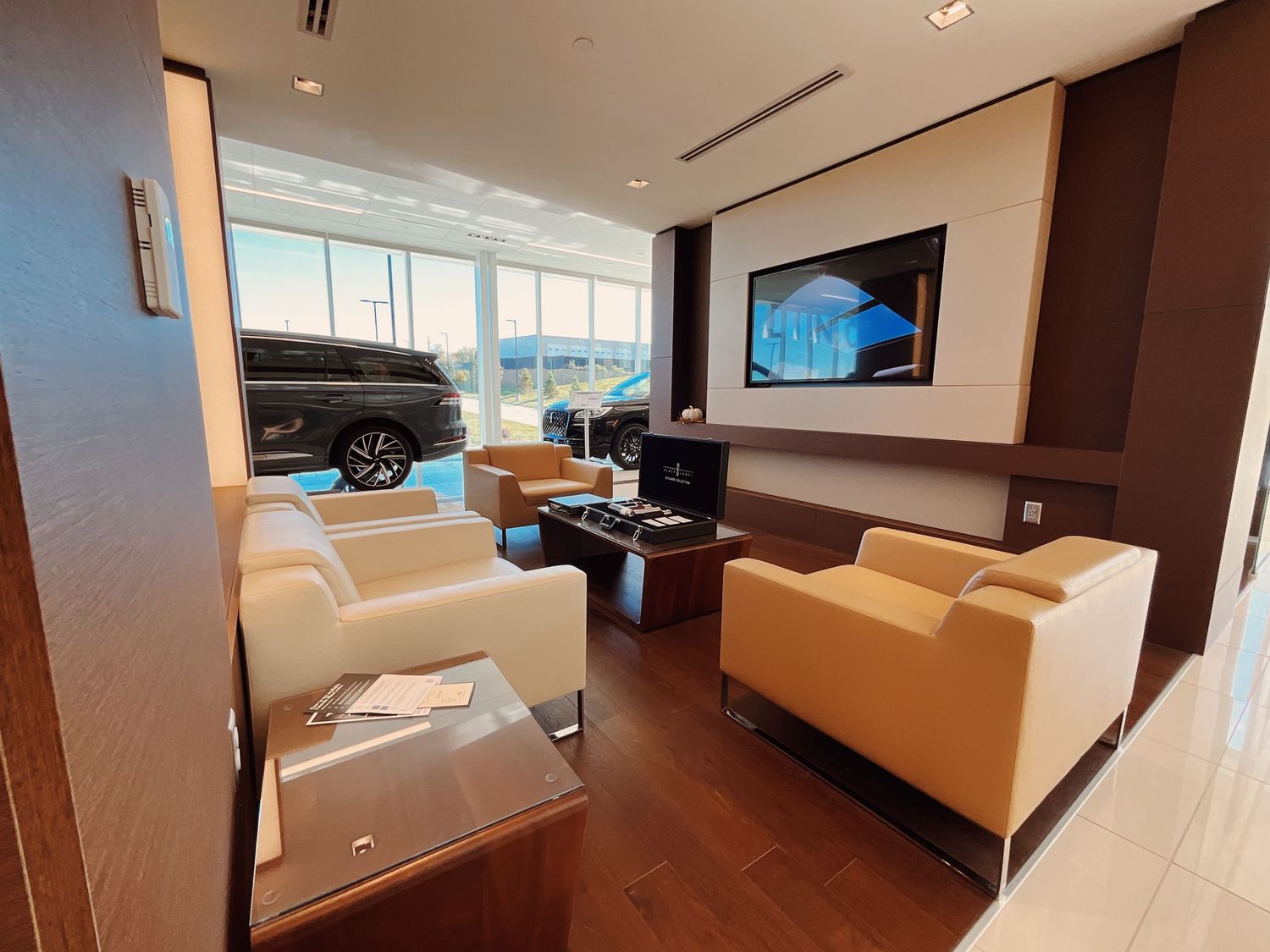 Modern car dealership lounge with cream sofas, dark wood floors, large TV display, and SUV visible through showroom window.