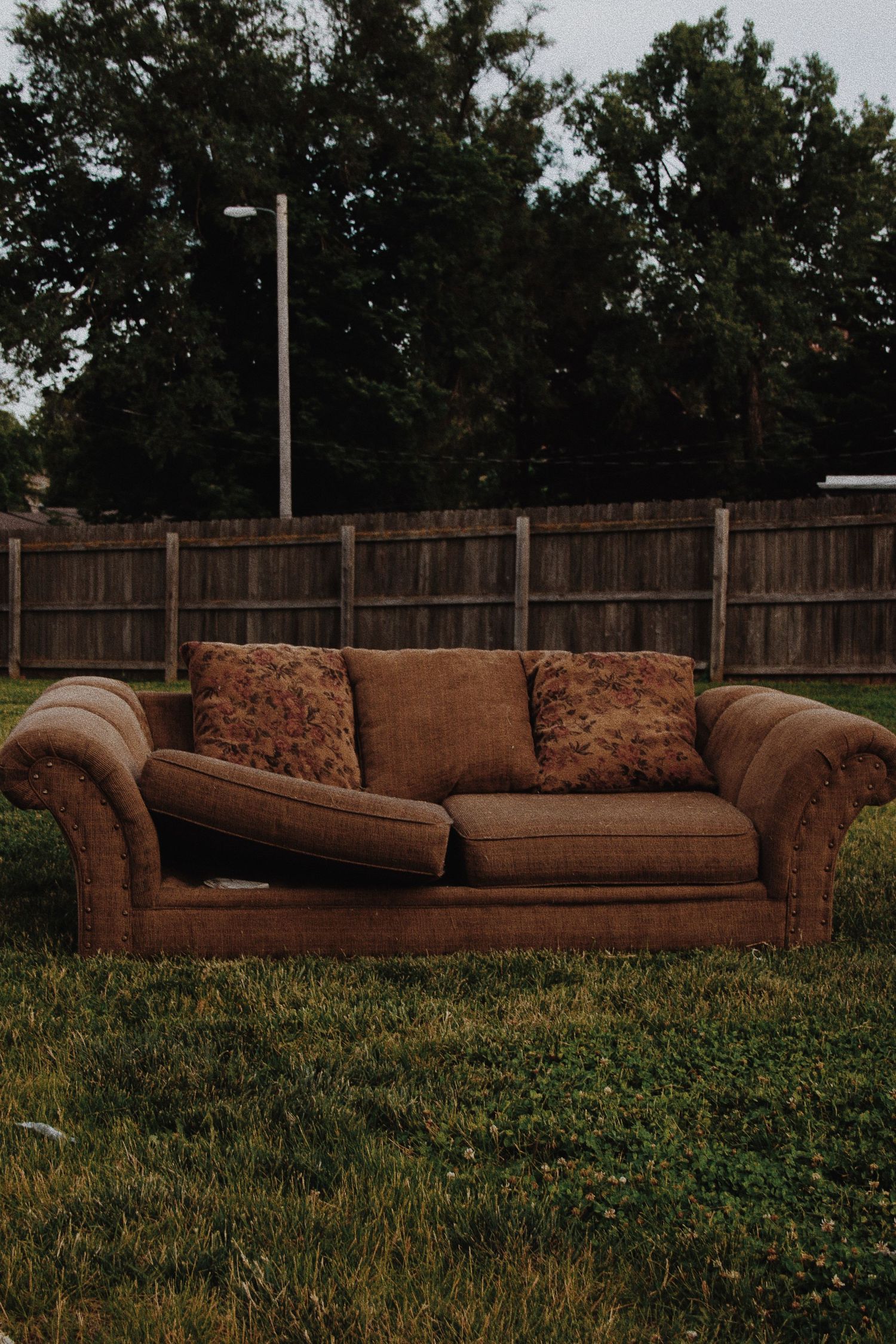 A worn brown sofa with floral pillows sits outdoors on green grass in front of a wooden fence.