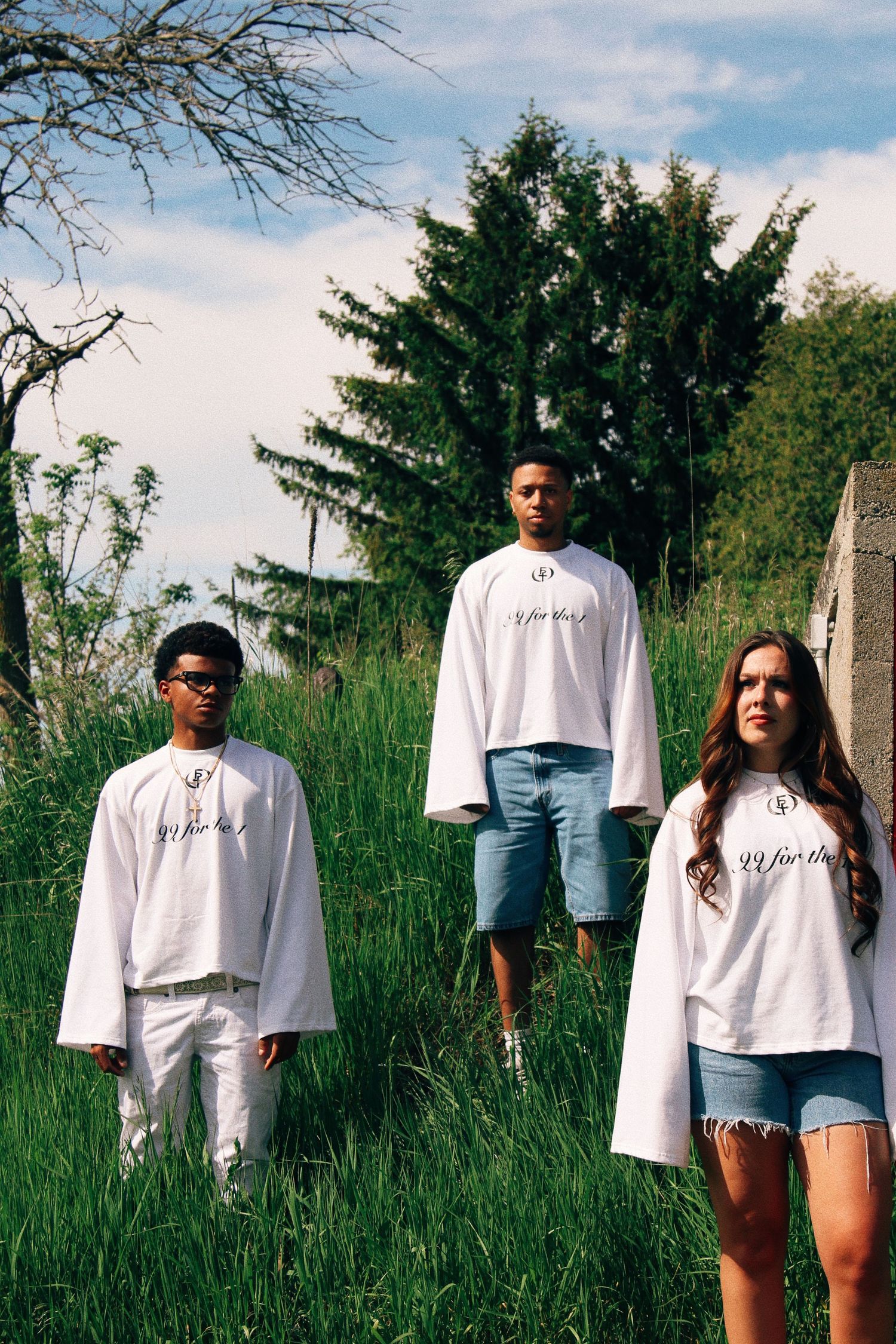 Three young people wearing white oversized graphic tees pose outdoors on a grassy hill with trees in background.