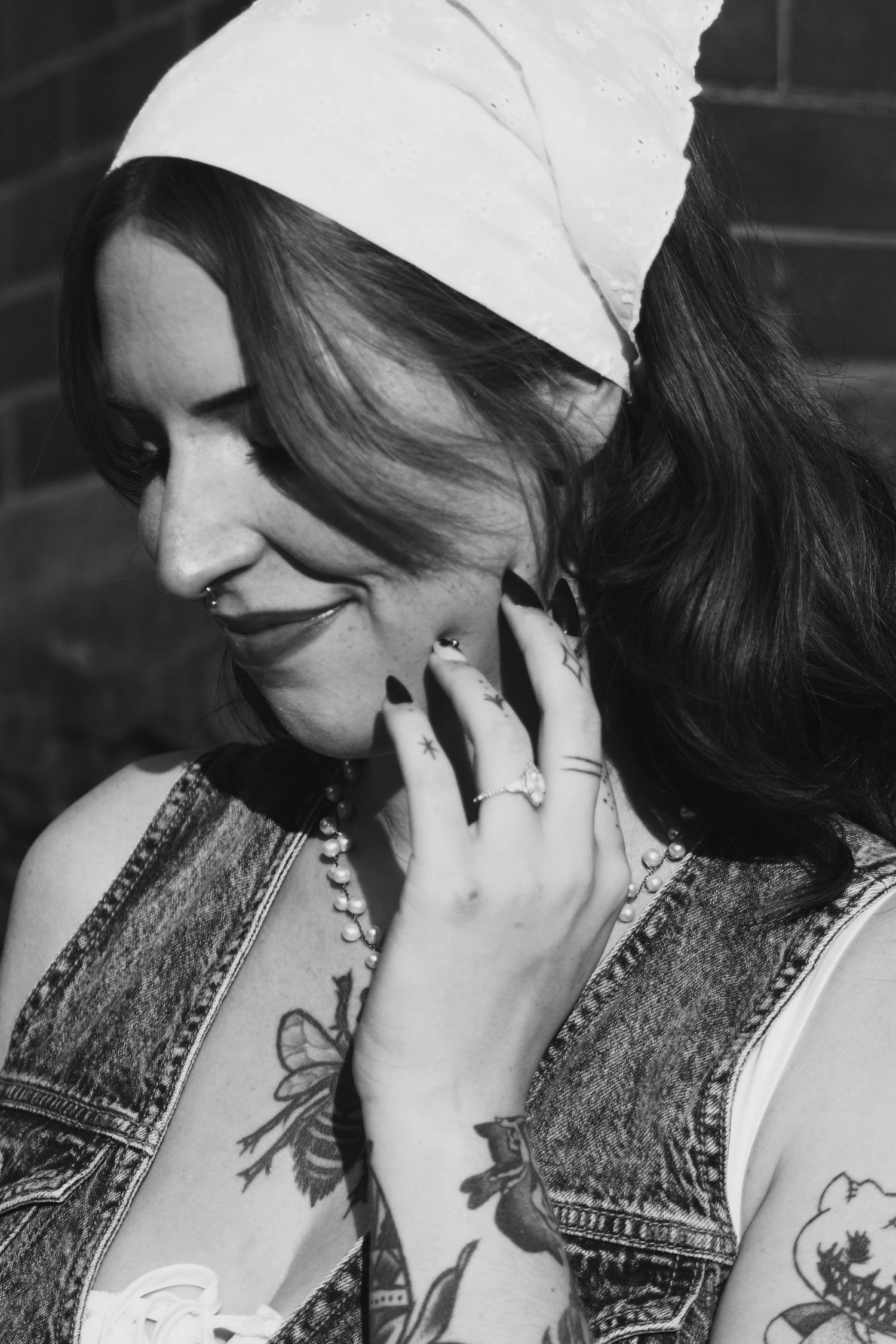 Black and white photo of tattooed woman with white headscarf, dark nails, and nose ring smiling softly.