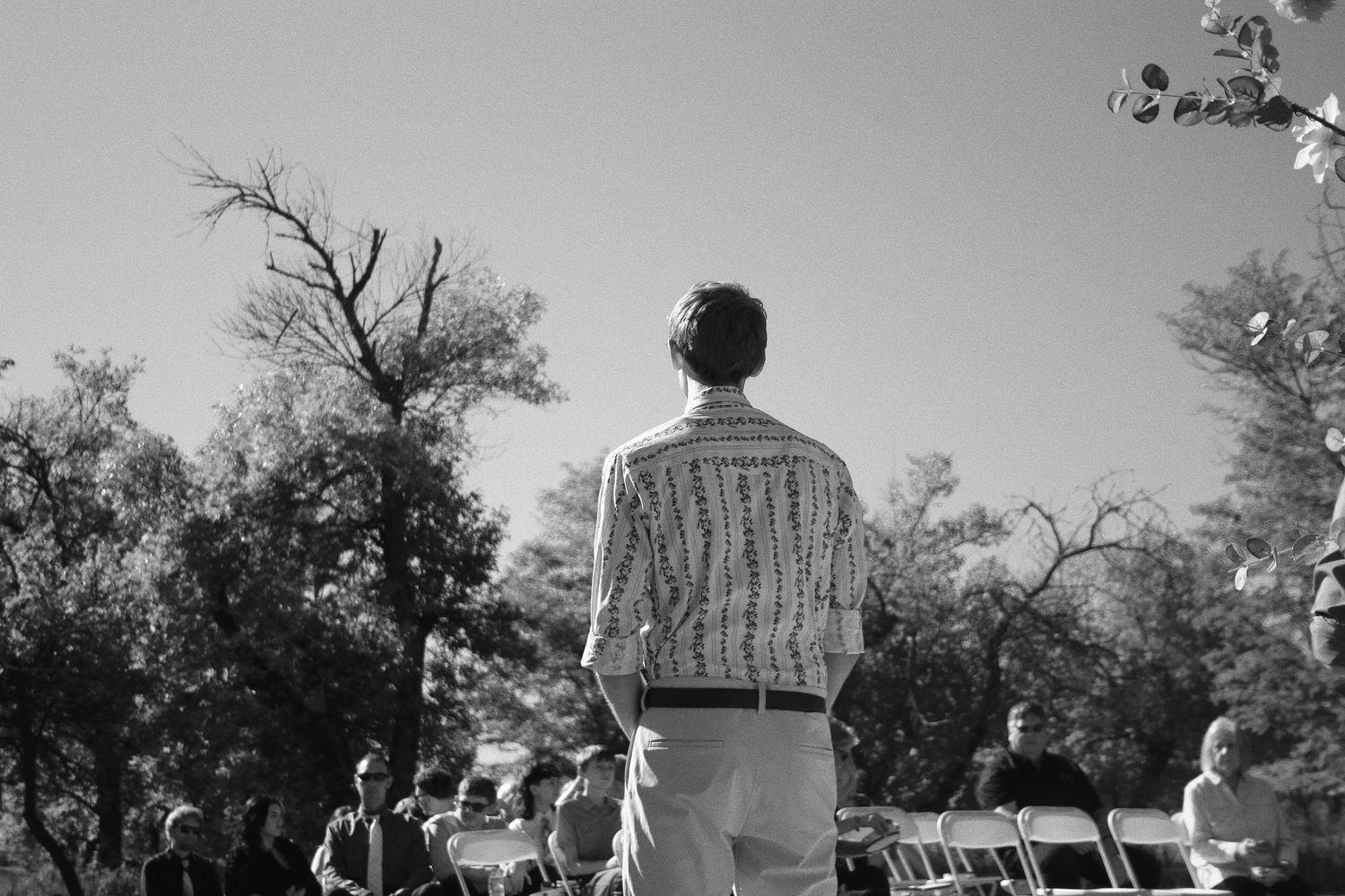 A person in a striped shirt stands outdoors at a gathering surrounded by trees and white chairs.